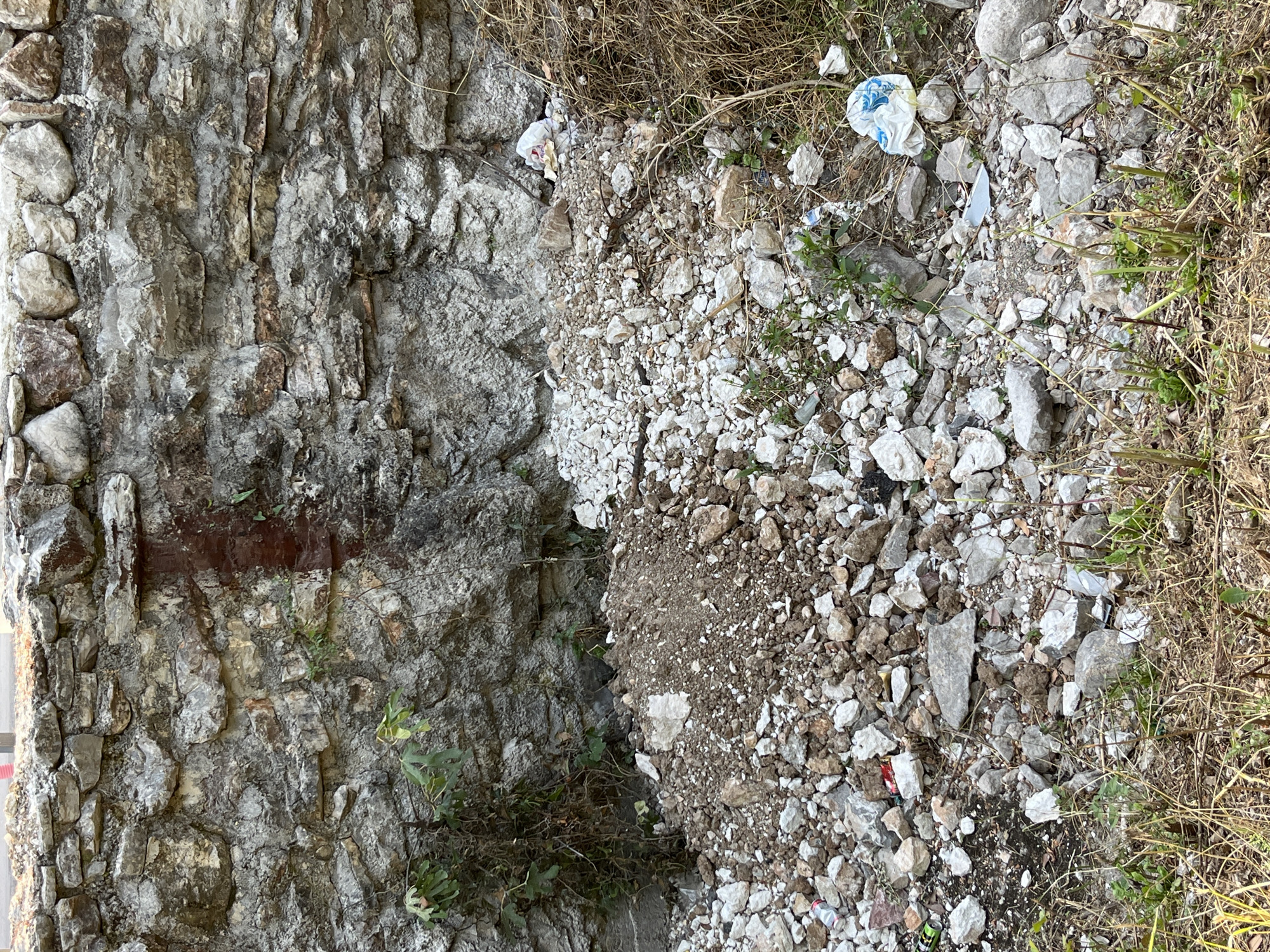 The image depicts a pile of rubble and debris in front of a stone wall. The wall appears to be constructed from irregular stones of varying sizes and colors, with some evidence of mortar. There is a reddish-brown stain running down a section of the wall. The ground in front of the wall is covered with a mixture of broken stones, dirt, and dry grass. Several pieces of white plastic trash bags are visible among the debris, as well as some scattered bits of paper and what appear to be metal cans. Small green plants are growing amongst the rubble and along the base of the wall. The scene suggests an area that has undergone demolition or construction, or is a neglected space accumulating waste. The location is identified as Budva, Montenegro. There are no people or animals visible in the image.