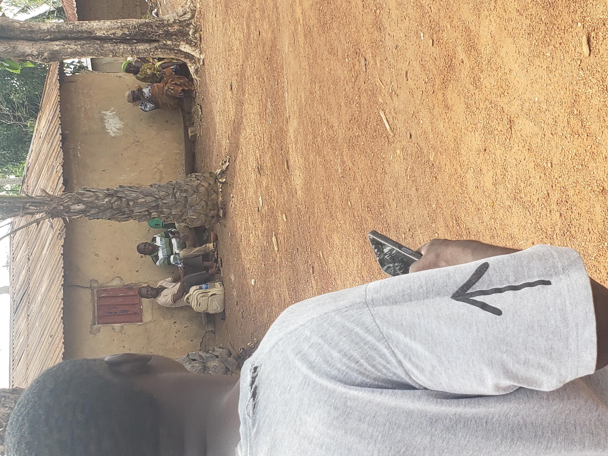 A medium shot captures a scene in the city of Kachia, Nigeria. In the foreground, the back of a person wearing a grey t-shirt with a black arrow drawn on the sleeve and a phone in their hand is visible. The t-shirt appears to have a tear near the shoulder. In the background, a simple, mud-brick building with a corrugated metal roof stands. Two men are seated on the ground in front of the building, one wearing a light-colored polo shirt and the other a striped green and white shirt. A light-colored backpack rests beside them. Further to the right, two women are also seated on the ground, one in a brown and white patterned skirt and the other in a green headscarf and yellow and brown patterned dress. They appear to be conversing. The ground is covered in red dirt. Trees are present, including a palm tree to the left and two large trees on the right.