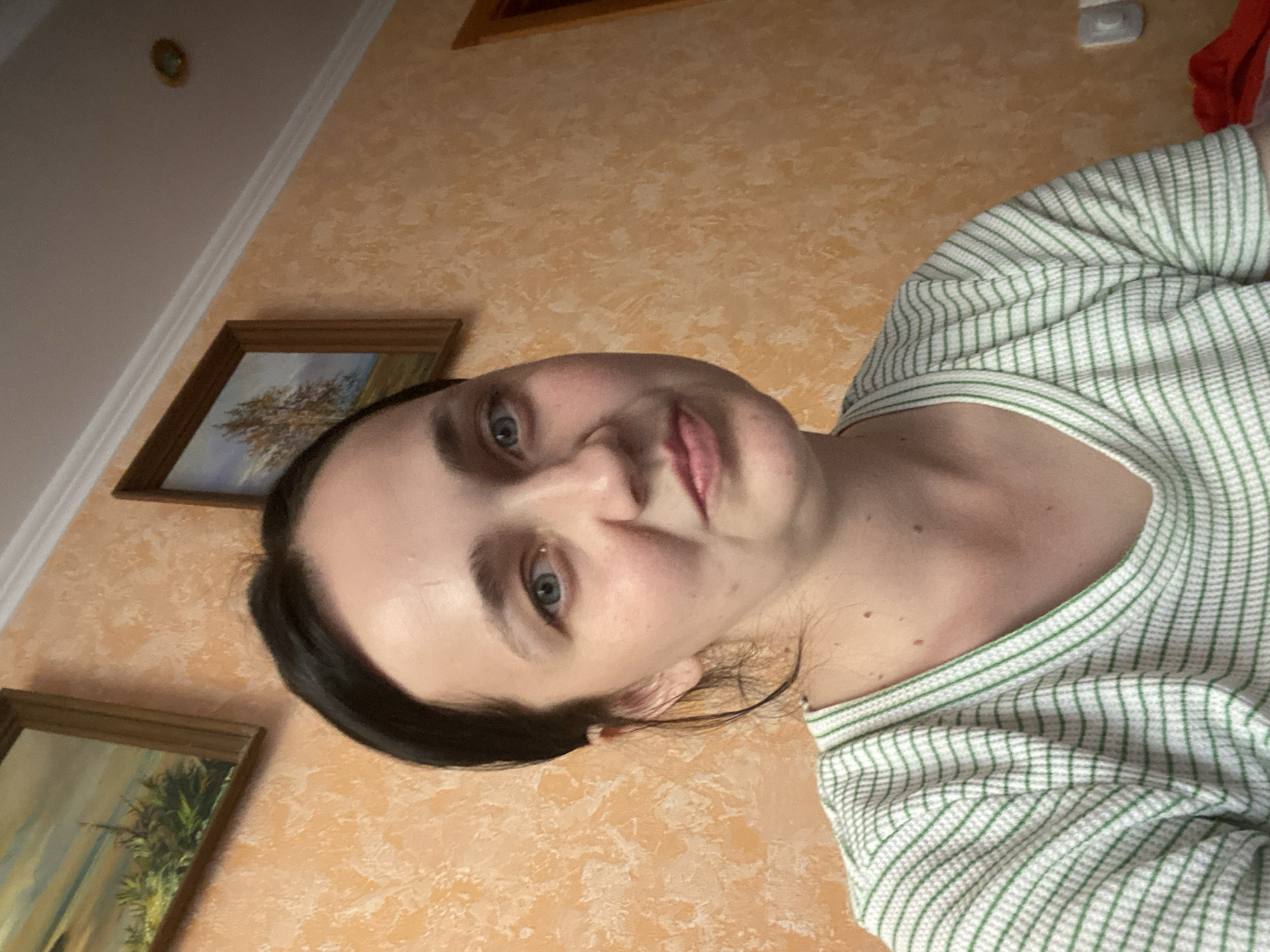 A medium shot shows a woman from the chest up, facing slightly to her right. She has dark hair pulled back and light blue eyes. She is wearing a green and white striped shirt. The background is an orange textured wall with two framed pictures hanging on it. The overall lighting suggests an indoor setting. There are no indications of a specific city or location.