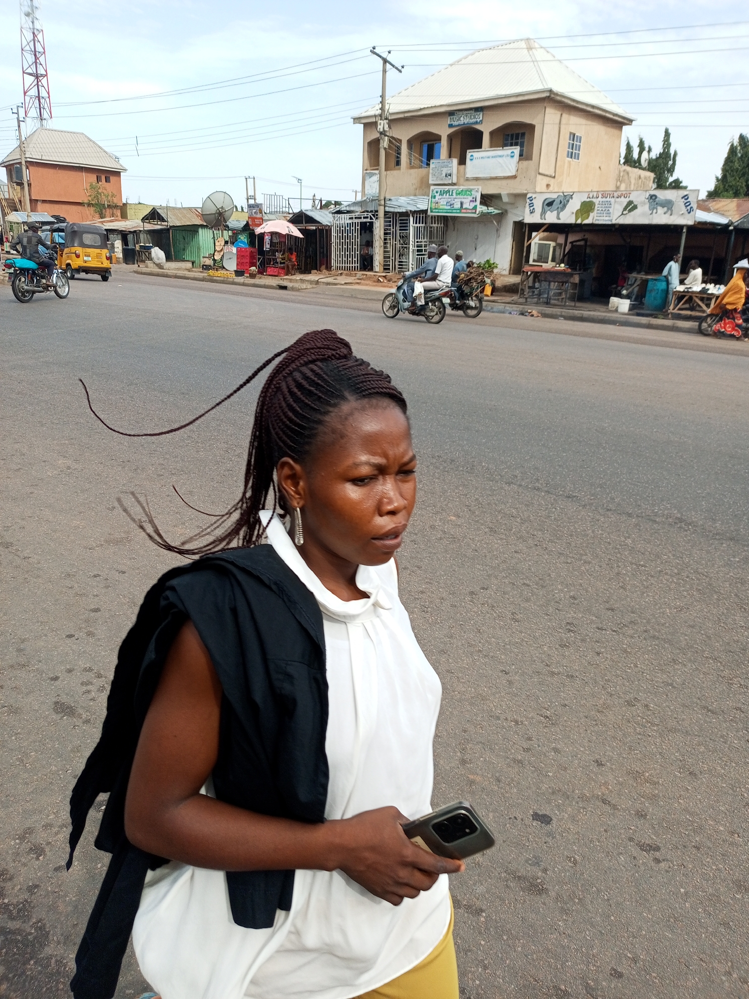 This image depicts a street scene in Tumfure, Nigeria. The central subject is a young Black woman with her hair in intricate braids, some of which are loose and trailing in the breeze. She is wearing a white top with a black garment draped over her shoulders and yellow pants. In her right hand, she holds a smartphone. Her expression is one of mild concern or focus.
The background reveals a typical street in a developing urban area. Several buildings line the road, some appearing to be shops with awnings. One building, taller than the others, has a light-colored facade and a prominent white roof. Signs are visible on some of the structures, including one for "Apple Drugs" and another with text in a local script, possibly indicating a food vendor such as "A.I.D SUYA SPOT." A large satellite dish is also visible.
Several vehicles are present on the road and roadside, including motorcycles, a yellow trishaw (auto-rickshaw), and a blue motorcycle with a rider. People are seen on and around the vehicles and in front of the shops. The time of day appears to be late morning or early afternoon, given the daylight and the angle of the sun, casting moderate shadows. The weather seems overcast, suggesting a possibly humid or temperate climate. The overall scene conveys a sense of everyday life and commerce on a Nigerian street.