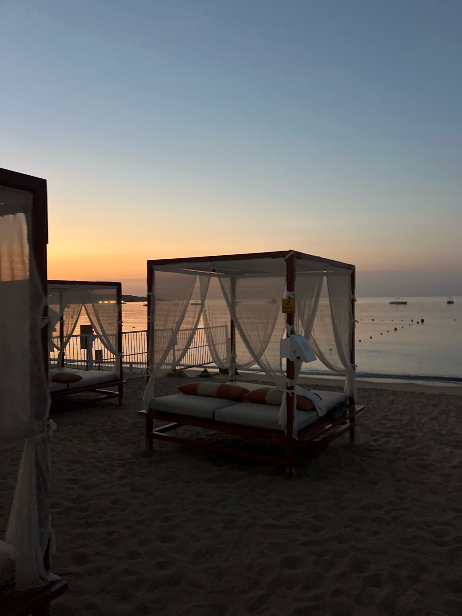 The image depicts a tranquil beach scene at dusk in Sharm El Sheikh, Egypt. The sky transitions from a soft blue at the top to a warm orange hue near the horizon, indicating the setting or rising sun. The foreground is dominated by sandy beach, with the shadows suggesting low light conditions. Two covered daybeds, typical of a resort setting, are prominently featured. These structures have wooden frames and are draped with white sheer curtains, with cushions visible on the beds. In the background, a calm body of water, likely the Red Sea, stretches to the horizon. A few boats are visible on the water, and a line of buoys marks a boundary. A metal railing runs along the edge of the beach, separating it from the water. The overall atmosphere is one of peacefulness and relaxation, characteristic of a beach resort.
