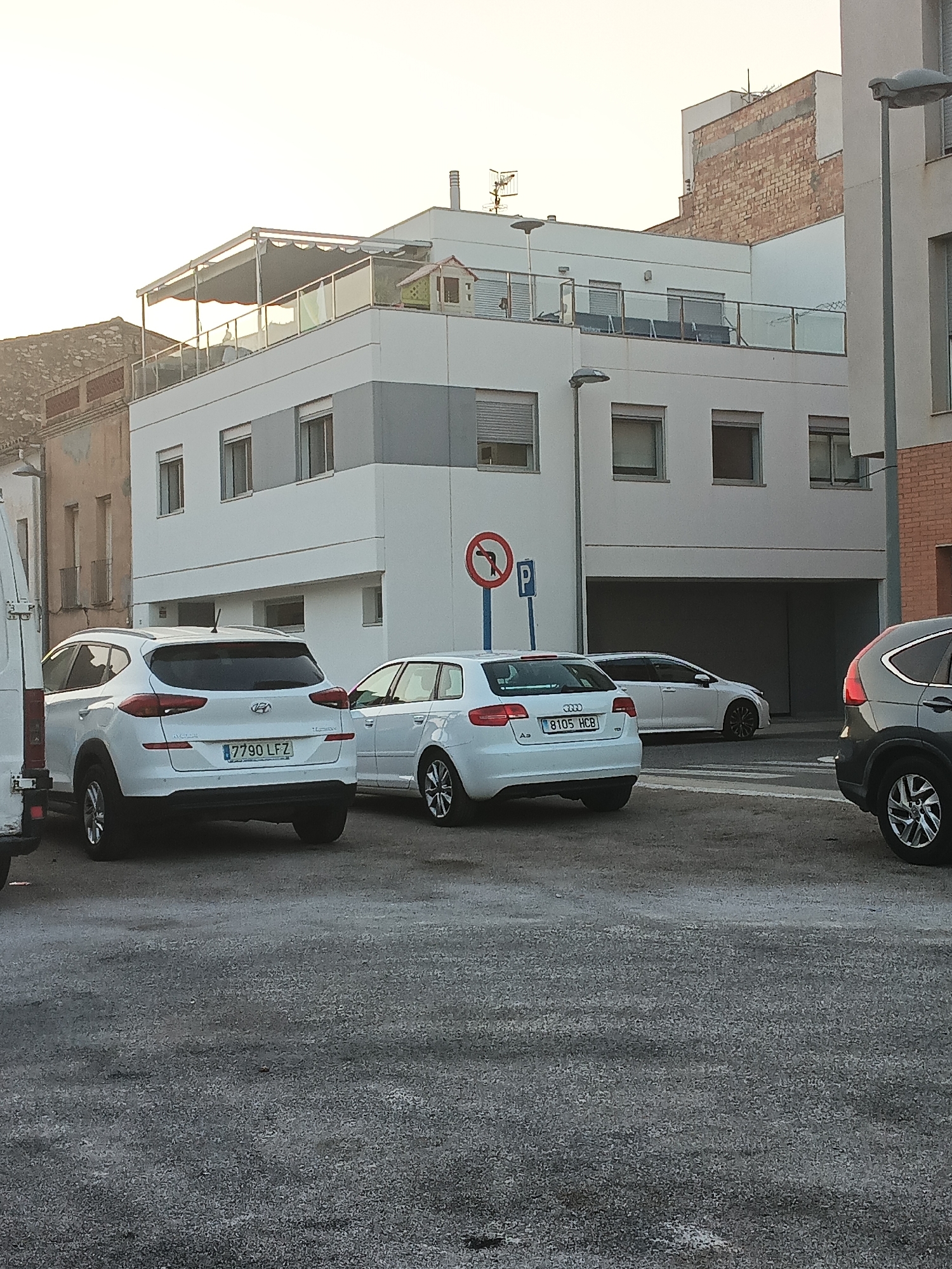 A white Hyundai Tucson SUV, license plate 7790 LFZ, is parked on an asphalt surface in front of a modern white building with multiple windows and a balcony on the second story. To the right of the Hyundai, a white Audi A3 hatchback, license plate 8105 HCB, is parked. A dark-colored SUV is partially visible on the far right. In the background, the white building has a darker gray band across its facade and a covered balcony with glass railings. A red and blue traffic sign with a red circle and a left-turning arrow with a red line through it, along with a blue "P" sign for parking, are visible on a pole. The sky is bright and appears to be either early morning or late afternoon. The scene is likely located in Tortosa, Spain, given the Spanish license plates and architectural style.