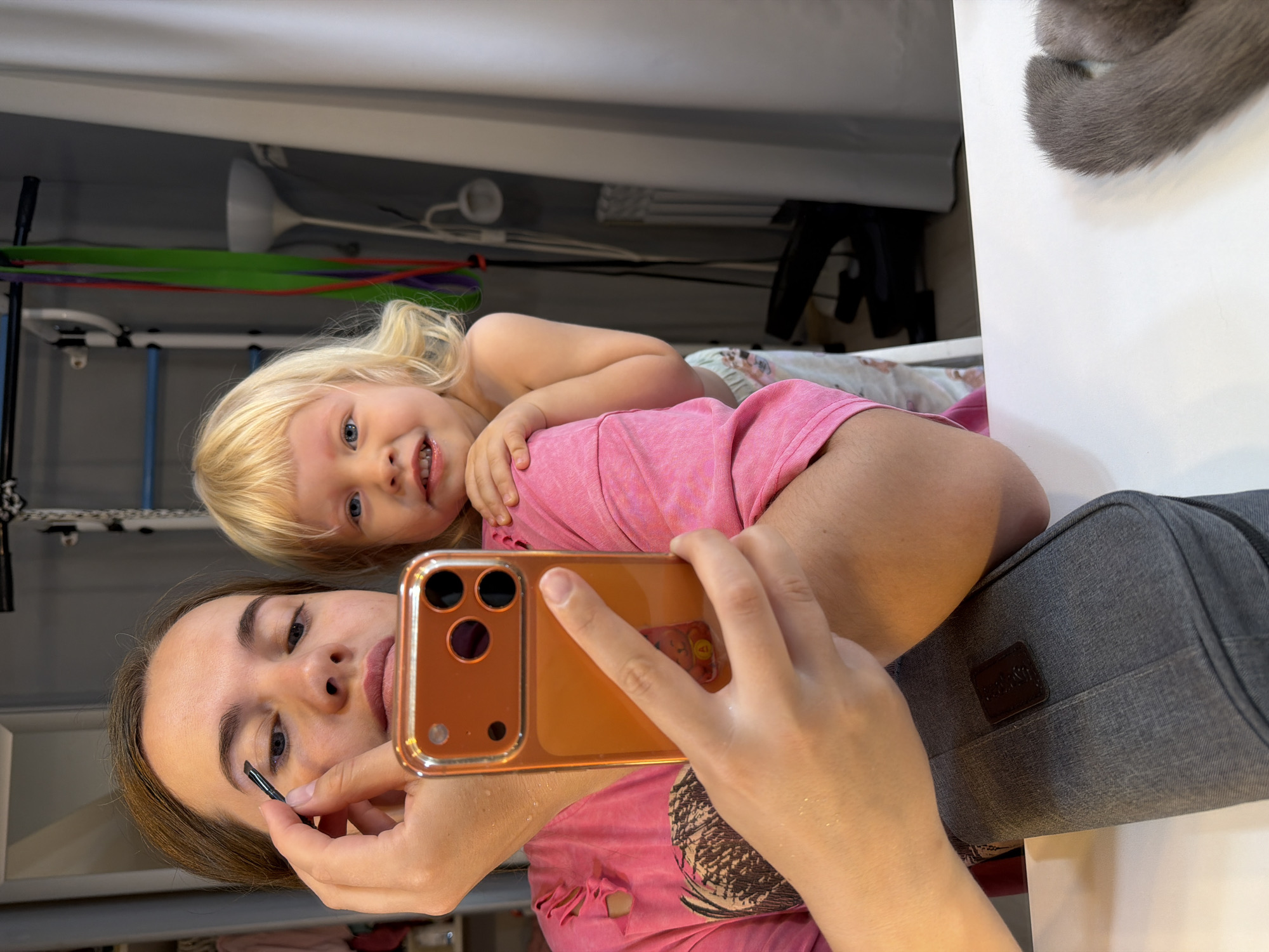A selfie shows a young woman and a blonde toddler, presumably a parent and child, lying down together. The woman is applying makeup, specifically to her eyebrows, while holding up a smartphone with an orange case. The toddler is smiling and snuggling close to the woman. They appear to be indoors in a home environment. Various pieces of furniture, workout equipment, and storage units are visible in the background. An animal (likely a cat) is visible in the top right corner, close to the ceiling. The exact location in Sochi, Russia, cannot be determined from this image.