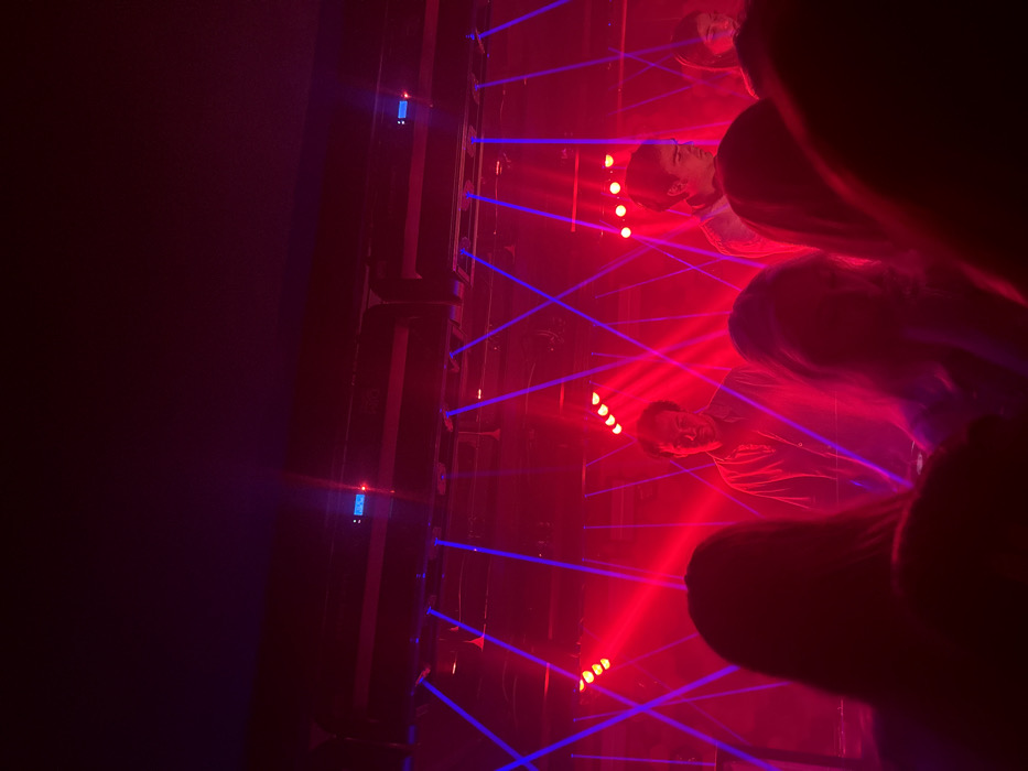 The image portrays an indoor music venue or club in Madrid, Spain. The specific location within Madrid cannot be identified from the visual information. The scene is enveloped in intense artificial lighting, predominantly red and blue.
In the immediate foreground, the backs of several individuals, presumably audience members, are visible as blurred silhouettes, facing towards a stage area. One person on the left has visible hair.
On the illuminated stage, two prominent male figures are present. The man on the left has dark, slightly curly hair, wears a collared garment, and is looking forward with a faint smile. The man on the right has dark, short hair, wears a light-colored top, and is looking downwards and to his right. A third, partially obscured figure is visible on the far right of the stage.
The environment above the performers is filled with stage lighting. Multiple small, circular red spotlights are arranged across the stage's width, casting a strong red glow. Numerous blue laser beams crisscross diagonally, creating a dense pattern across the upper and middle portions of the scene. The ceiling structure of the venue is dark and visible in the upper frame. The atmosphere appears to contain haze or smoke, making the light beams more distinct.