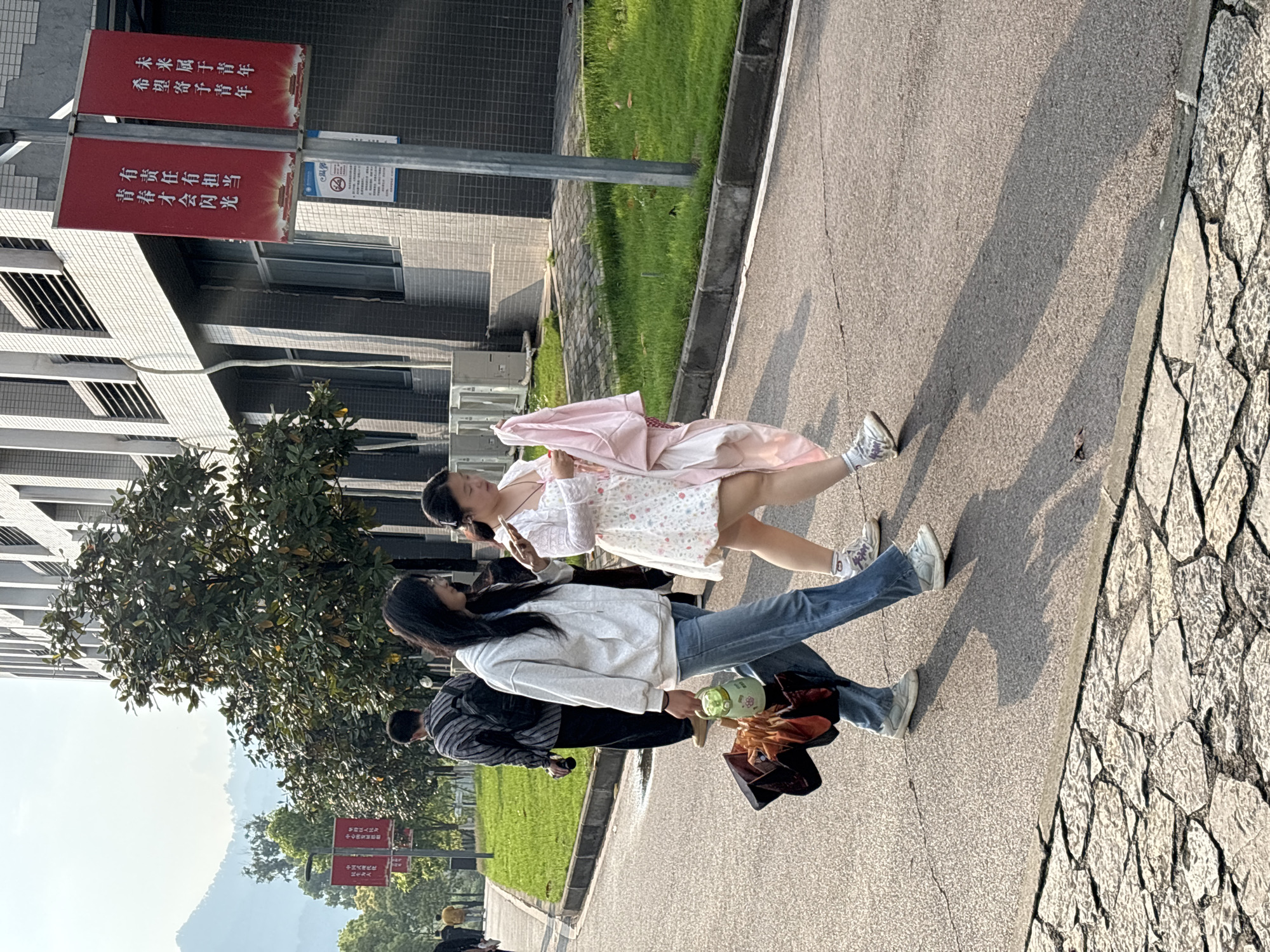 Two women are walking along a paved walkway in Jiangjin, China. The woman on the right is wearing a short, floral-patterned dress with a pink jacket draped over her arm. She is looking at a cell phone. The woman on the left is wearing jeans, a white hoodie, and carrying a green water bottle and a brown bag. A man is visible in the background, walking behind them. A building with many windows is visible on the left side of the image. Two red signs with white Chinese characters are attached to a pole in front of the building. One sign reads, "Hope is entrusted to the youth," and the other, "The future belongs to the youth." Mountains are visible in the distance.
