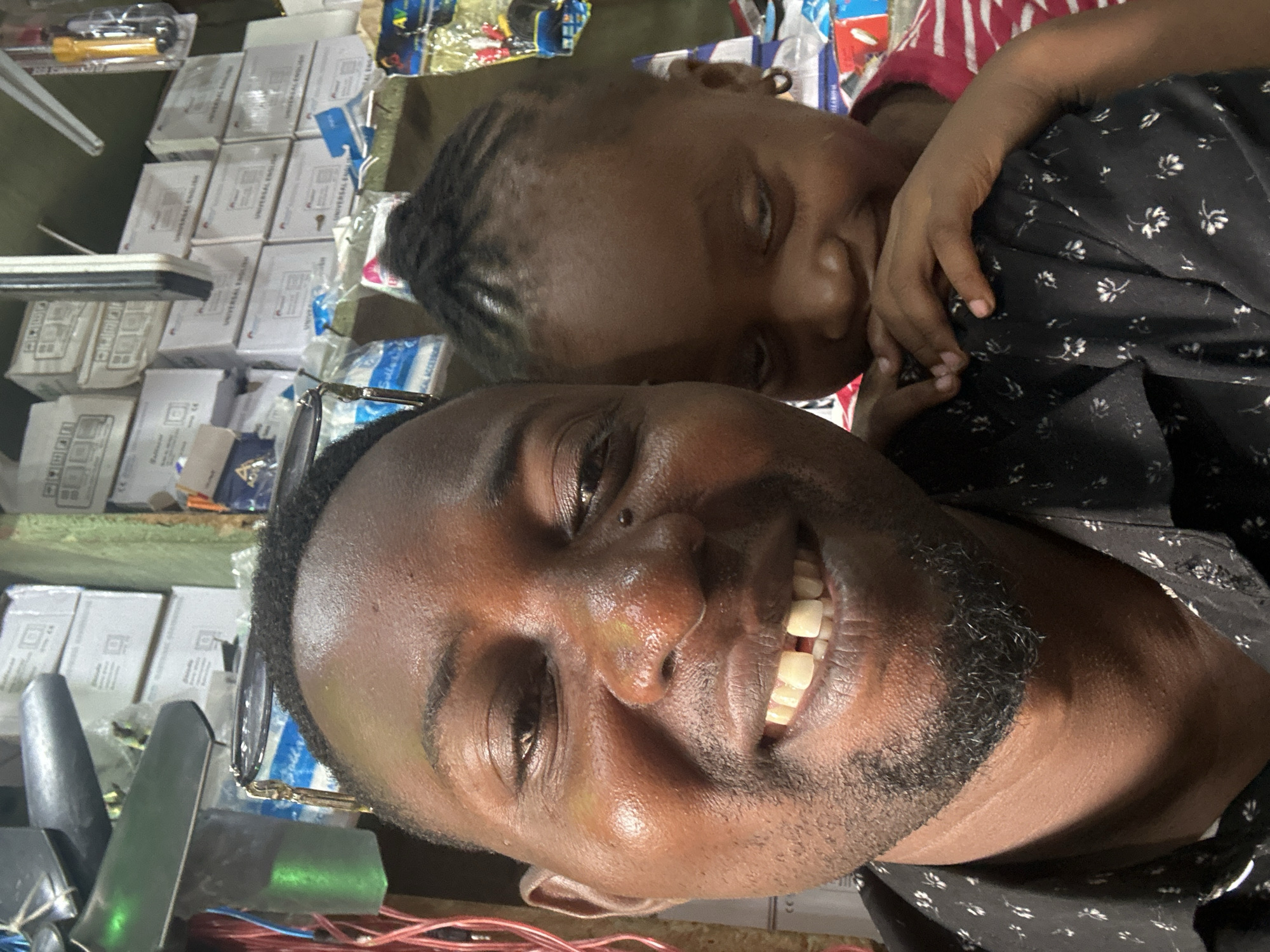 The image captures a close-up, selfie-style photograph of a man and a young child in what appears to be a shop in Narayi, Nigeria. The man, on the left, is smiling broadly, revealing a gap in his front teeth. He is wearing a dark shirt with a white floral pattern. His skin appears to be a medium to dark brown. The child, on the right, is leaning into the man, with their face partially obscured by their hands. The child has dark hair styled in small braids and is wearing a pink and white striped shirt.
The background of the image is cluttered with various items, suggesting a retail environment. Cardboard boxes, some stacked on shelves, are visible. There are also some wires and possibly electrical components, including a green-colored object that resembles a light or fixture. The overall impression is of a busy, perhaps small, store. The lighting suggests it is daytime, with some artificial light also present. The image is taken from a low angle, emphasizing the faces of the man and child.
