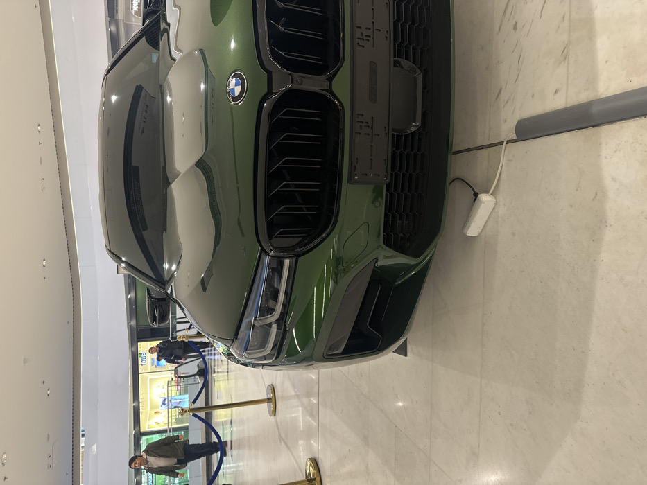The image displays the front section of a dark green BMW sedan, positioned as a display vehicle inside a modern shopping mall. The car features a prominent black kidney grille, LED headlights, and a black placeholder license plate holder on its front bumper. A white power strip with a cable is visible on the light-colored tiled floor, extending from under the car to a grey cable management strip. In the background, behind gold stanchions and blue ropes, two individuals are seen walking away from the camera. Retail storefronts, including one for "TOUS," line the mall corridor, and an escalator is visible in the distance. The setting suggests a high-end commercial environment, likely Tashkent City Mall in Tashkent, Uzbekistan, given the architecture and brand presence.
