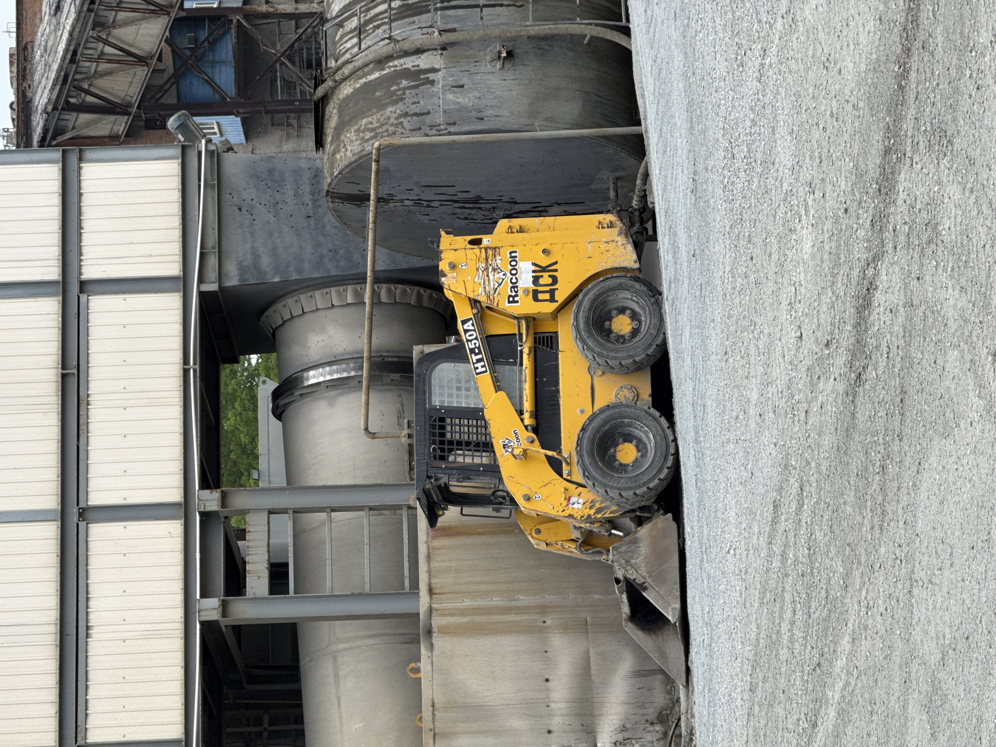 An industrial scene captured in Vladivostok, Russia, features a yellow skid-steer loader positioned among large industrial structures and a significant pile of granular material.
The primary subject is a yellow skid-steer loader, appearing to be an HT-50A model from a brand called "Raccoon," indicated by text and a raccoon head logo on its side. Another piece of text, "ACK," is also visible on the machinery. The loader has a front bucket, four wheels, and a protective cage over the driver's cabin, suggesting heavy-duty use. It is positioned next to a massive, light grey pile of granular material, likely aggregate, sand, or gravel, which dominates the right side of the frame.
In the background, a complex industrial facility is visible, featuring multiple large, grey cylindrical silos or tanks, varying in size and height. These structures are connected by pipes and supported by a robust framework of steel beams and girders. Parts of a building with corrugated metal siding are also seen on the left, blending into the industrial environment. The lighting suggests it is daytime, possibly under an overcast sky, as there are no strong shadows. No people are visible, emphasizing the mechanical and functional nature of the setting. The scene conveys the activity of material handling within a plant or construction site.