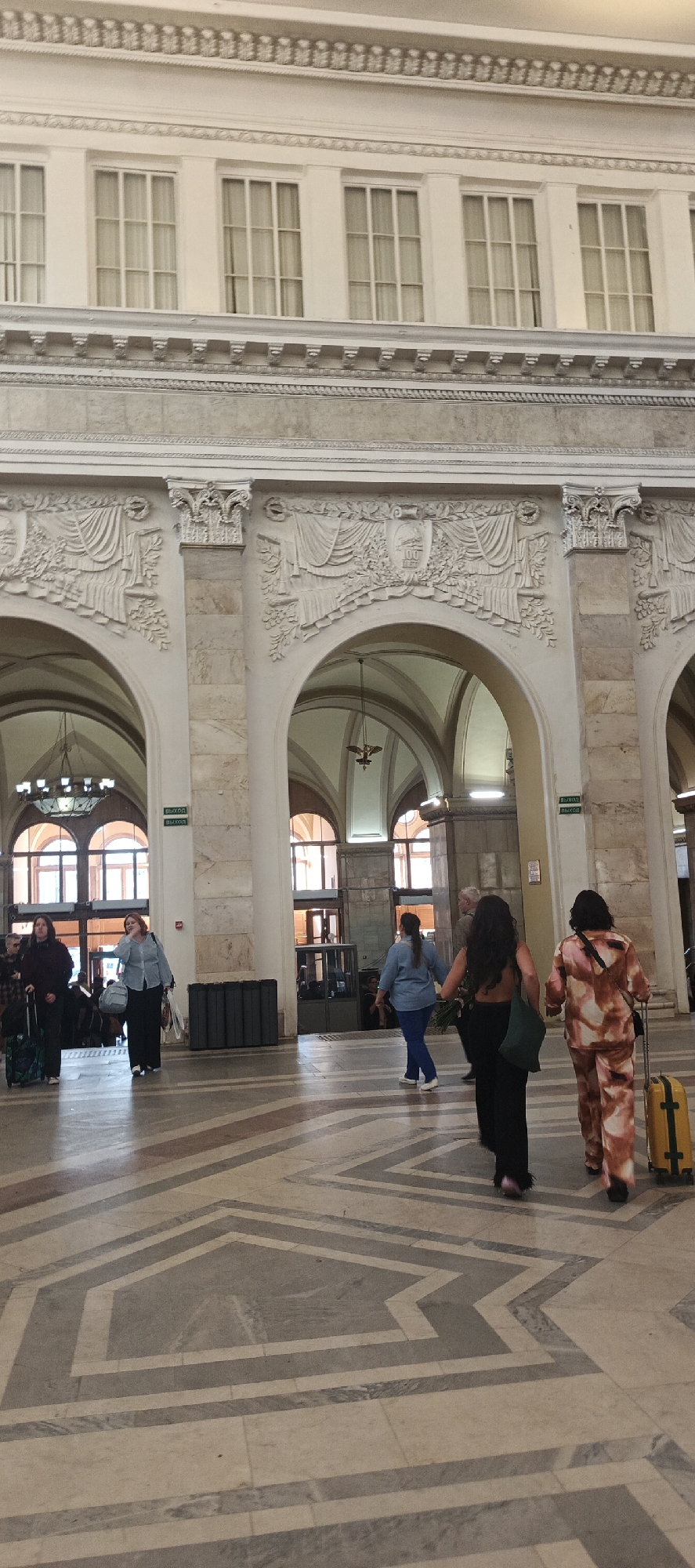 The image depicts the interior of a grand railway station, likely the Vitebsky Railway Station in Saint Petersburg, Russia, given its architectural style. Multiple arched passageways, characteristic of 19th and early 20th-century European train stations, dominate the scene. The station features high ceilings, ornate decorative elements above the arches, and large, multi-paned windows that suggest ample natural light. The floor is paved with a geometric pattern of marble or stone tiles in shades of beige, grey, and white.
In the foreground and midground, several individuals are visible, suggesting moderate foot traffic. On the left, a woman in a dark top and pants, pulling a patterned suitcase on wheels, walks away from the camera. Next to her, a woman in a light blue denim shirt and dark pants stands, looking to her left and touching her face. Further into the station, two women are walking away from the camera. One wears a dark sleeveless top and black wide-leg pants, carrying a green tote bag. The other wears a patterned tracksuit in shades of pink and orange, pulling a yellow suitcase on wheels. In the background, more people can be seen, and another woman in a blue denim shirt and blue pants walks towards the right.
The environment is well-lit, with artificial lighting fixtures visible in the arched spaces. Green exit signs are strategically placed near the passageways. The overall impression is one of an active, historically significant public space.
