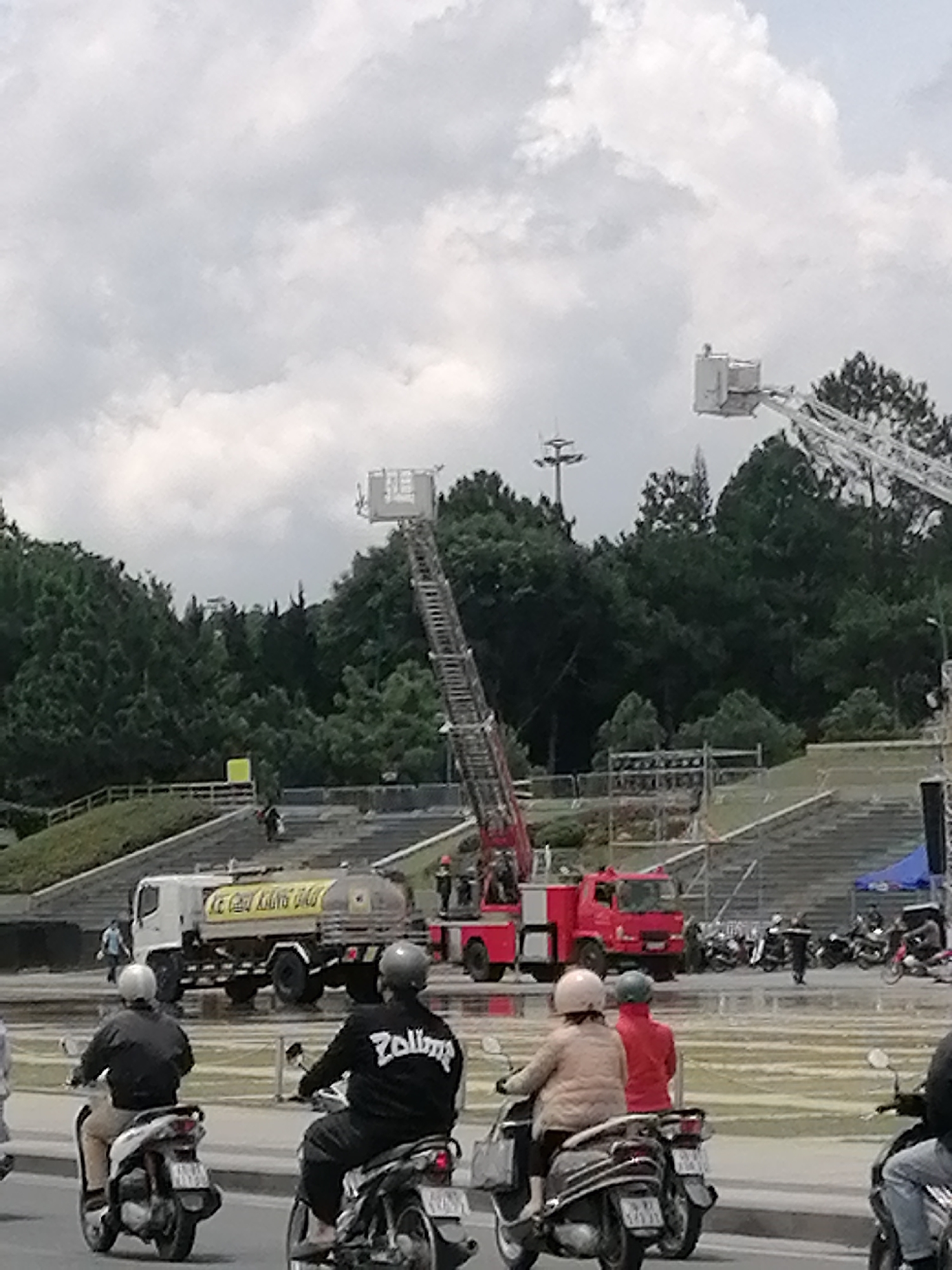 The image depicts a street scene in Da Lat, Vietnam, with a focus on a large white truck with a yellow advertisement on its side, a red fire truck with a ladder extended, and several people riding motorbikes. The truck appears to be a water truck. In the background, there are large green trees, a tiered structure that resembles stadium seating, and a cloudy sky. Several individuals are visible, some on motorbikes, others walking or standing near the vehicles. The overall impression is of a busy urban environment, possibly during an event or a public service operation.