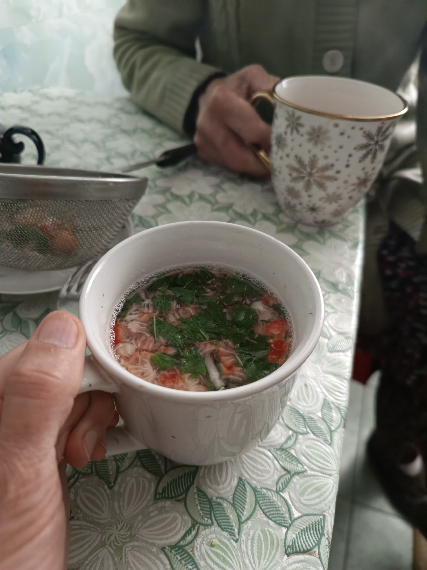 A close-up shot depicts a person holding a white mug filled with a clear broth containing shredded red meat, noodle-like strands, and chopped green herbs, possibly mint or parsley. Beside the mug, a metal mesh strainer rests on a small white plate, and a portion of its contents are visible. Another person is seated in the background holding a white and gold decorative mug. The scene is set on a table covered with a green and white floral patterned tablecloth. The location is Ostrogorka, Russia.