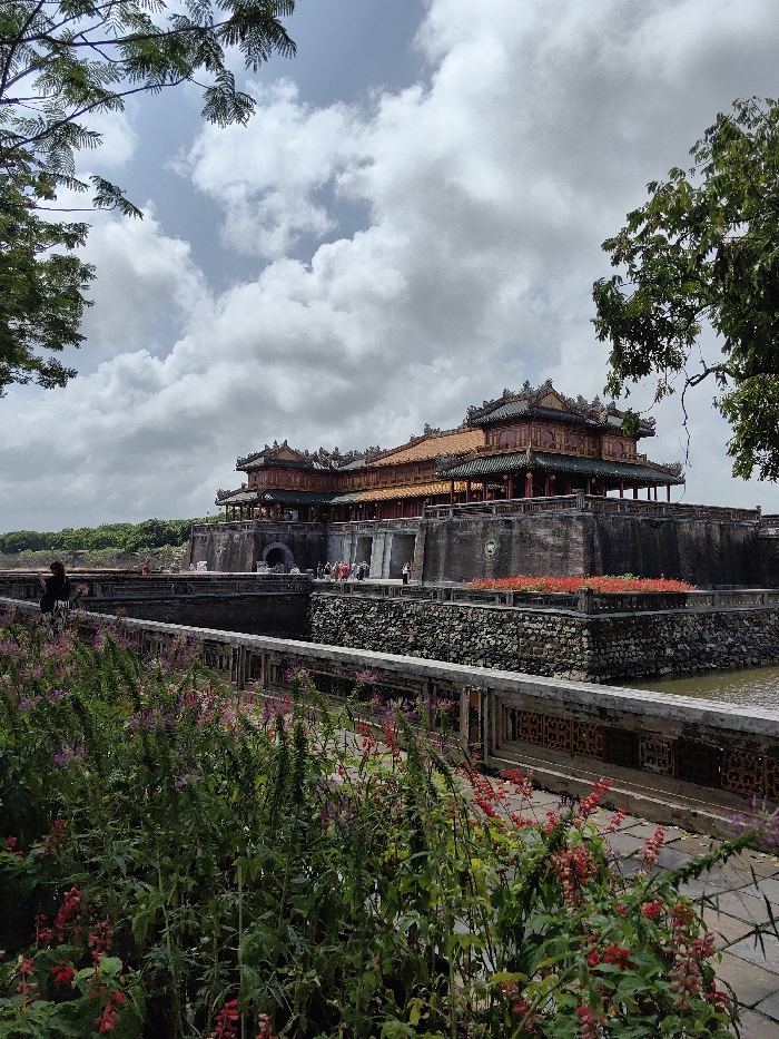 This image captures the Ngọ Môn Gate (Meridian Gate) of the Imperial City of Huế, located in Phú Xuân Ward, Vietnam, under an overcast sky with diffused light.
In the foreground, a dense bed of green plants with scattered red and purple flowers occupies the lower left. A light-colored concrete pathway with decorative balustrades runs horizontally across the mid-foreground, bordering a dark body of water, likely a moat. Beyond the moat, a robust, dark grey stone wall, constructed from stacked rough stones, forms a bank, featuring a rectangular planter box filled with vibrant red flowers in the mid-right.
The central architectural subject is the multi-tiered Ngọ Môn Gate, presented as a prominent structure built on a raised dark grey stone platform. The gate features traditional Vietnamese design elements, including elaborate dark-tiled roofs with decorative finials, visible red support columns, and lighter orange-ttiled upper sections. A large arched entrance is centrally positioned within the stone platform, with additional smaller arched or rectangular openings visible. Approximately 15-20 individuals, appearing to be visitors, are congregated on the platform and near the gate entrance. A single individual in dark clothing stands on a pathway to the far left, facing the gate.
A stone bridge crosses the moat, providing access to the gate's platform. Green tree foliage partially frames the image from the upper left and right corners. A continuous band of green trees lines the top of the distant wall, which extends horizontally behind the main gate structure.