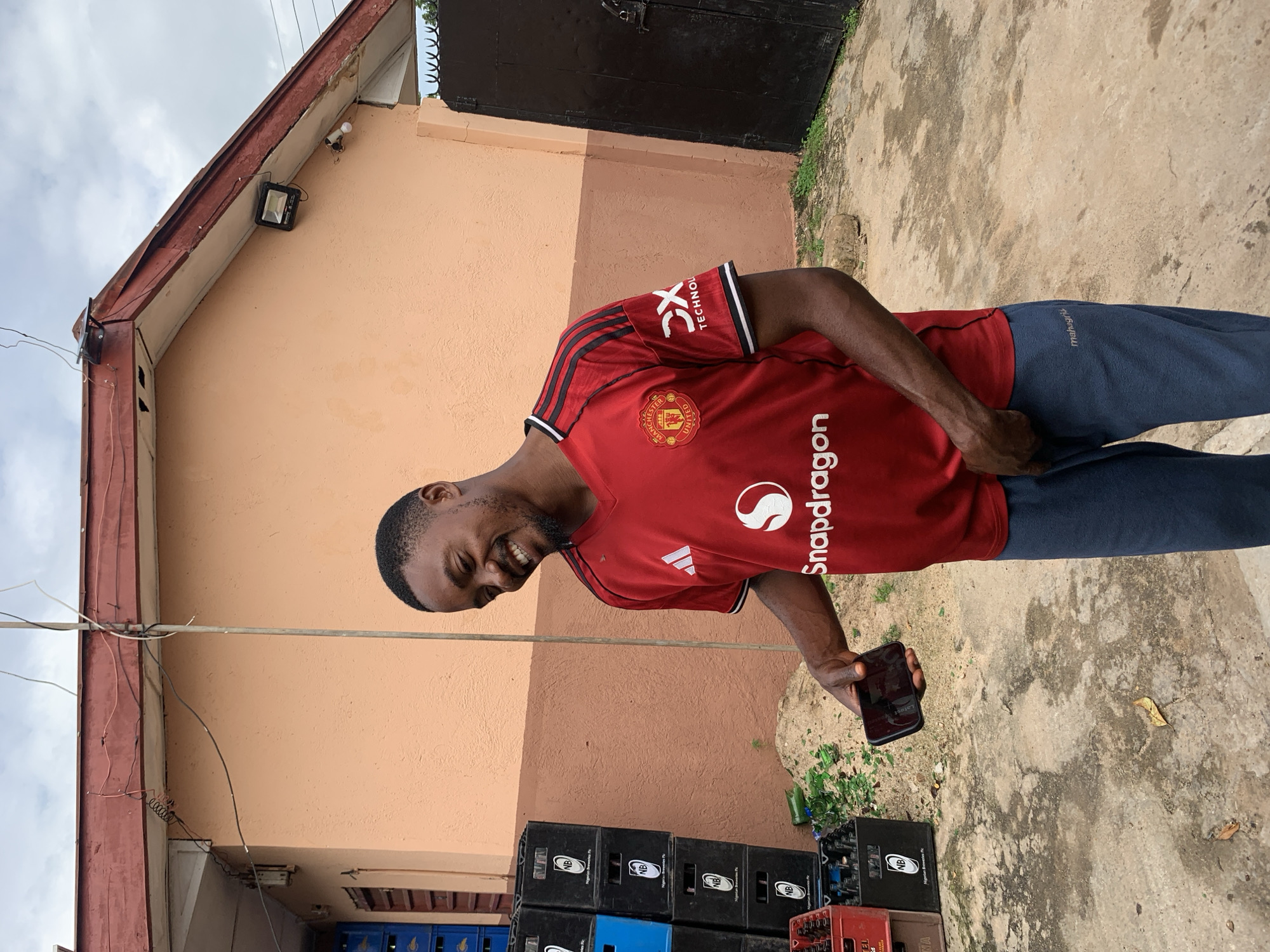 A medium shot captures a Black male in his 20s standing outdoors, holding a smartphone in his left hand, which is at his waist. He is wearing a red Manchester United jersey with black and white accents on the sleeves and collar. The jersey has a white Adidas logo on the left chest and the Manchester United crest on the right. The jersey also features a white "Snapdragon" logo on the front. He is also wearing dark blue sweatpants with a small white logo on the right leg that reads "mahagri.co". The man is looking to his right, with a wide smile and his head tilted back slightly.
The background consists of a light orange building with a dark red roof. Wires are strung across the front of the building and connected to a utility pole. To the left of the man, stacks of black plastic crates, some with a blue tint, are visible. Broken glass bottles are scattered on the ground near the crates. To the right of the man, a dark metal gate is visible. The ground is a rough, uneven concrete surface. The overall setting suggests an outdoor area, possibly a commercial or storage space, consistent with areas in Jos, Nigeria. The sky is overcast.