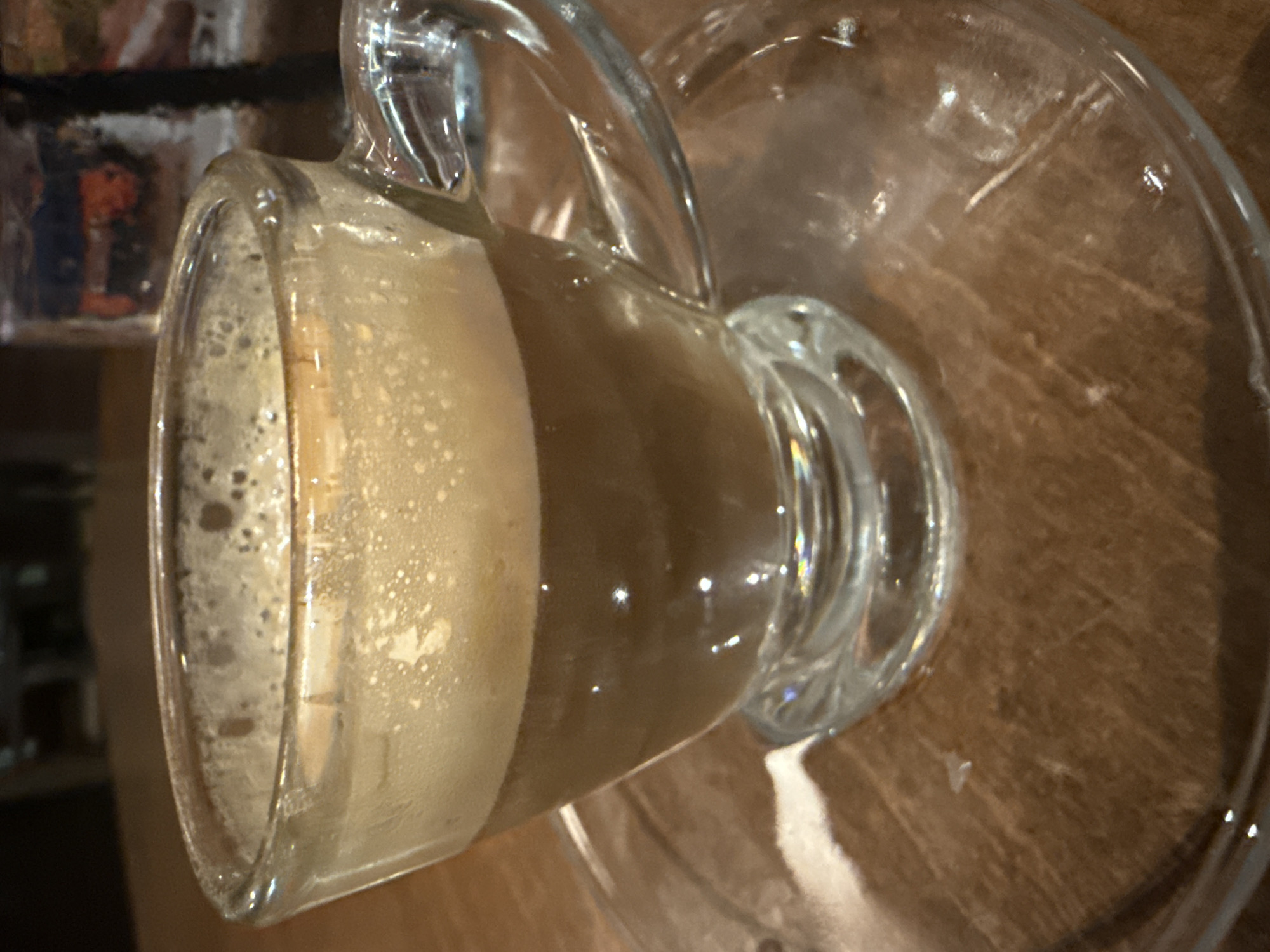 The image shows a glass mug with a handle, partially filled with a beverage, likely coffee or a similar foamy drink. The mug sits on a clear glass saucer placed on a wooden surface. The environment appears to be indoors. Though I do not have enough information to determine a specific location in Dania Beach.