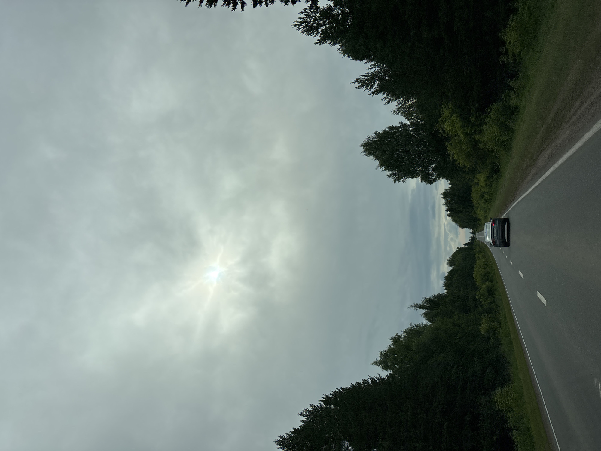 A two-lane asphalt road stretches into the distance, flanked by dense coniferous and deciduous trees. The sky is overcast with the sun partially obscured by clouds, creating a bright halo effect. A dark-colored sedan is traveling away from the viewer in the right lane. The road surface appears to be in good condition with white dashed lines marking the lanes. The environment suggests a rural or semi-rural setting, likely within the city of Забегалово, Russia, given the context.