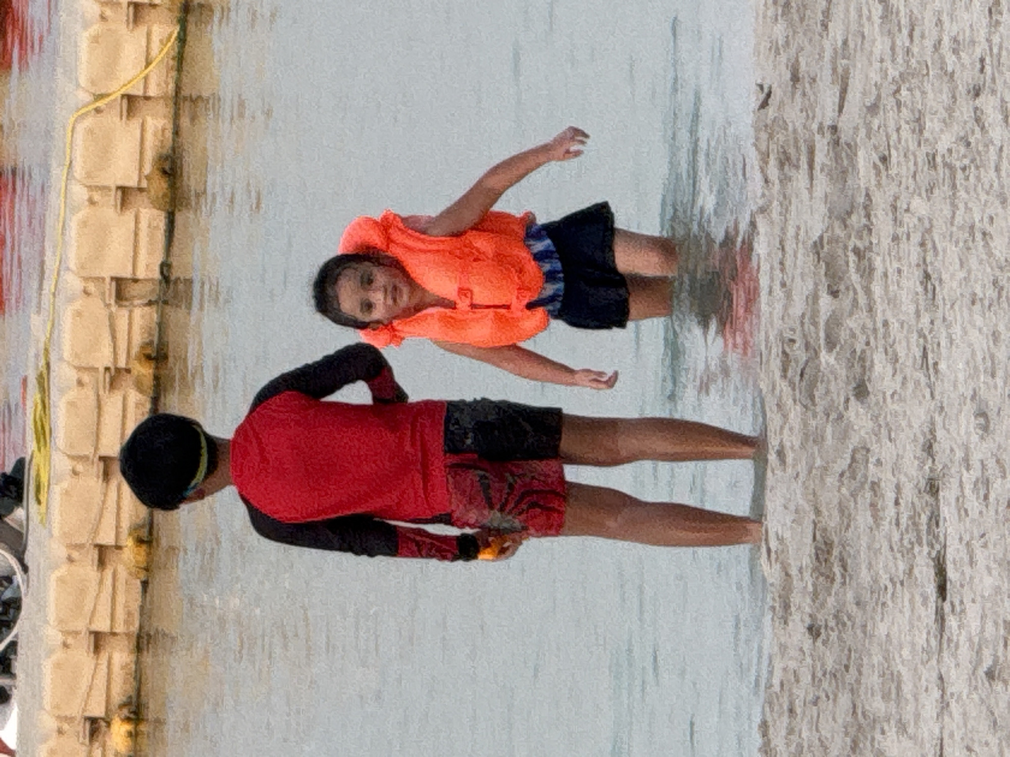 The image shows two children wading in shallow water at a beach. On the left is a boy wearing a red and black rash guard and patterned swim trunks. He has a black cap on and is facing away from the camera. On the right is a younger girl wearing an orange life vest over a blue and white striped swimsuit and dark shorts. She is facing the camera and appears to be looking slightly to her left. The water is calm and appears to be a light blue-gray color. In the background, there are yellow floating barriers and a yellow rope. The foreground consists of a sandy beach with some debris. The lighting suggests it is daytime. The location is specified as Unknown, United Arab Emirates.