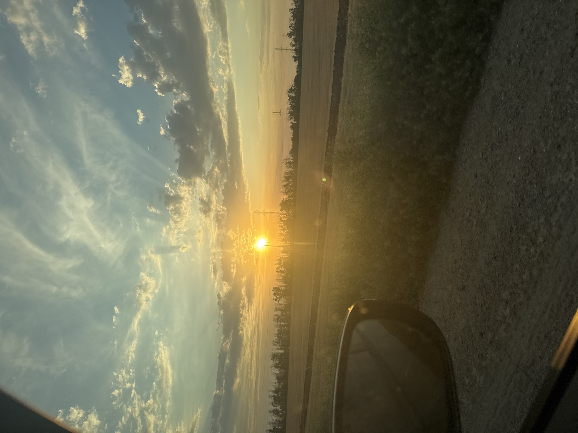 The image depicts a scenic view of a sunset over a rural landscape, likely in Taganrog City District, Russia. The sun, a bright orange orb, is setting behind a line of trees and power lines on the horizon. The sky is a canvas of colors, with streaks of white and gray clouds against a gradient of blue and yellow hues. In the foreground, a car's side mirror is visible on the left, reflecting the warm tones of the sunset. The terrain appears to be a mixture of fields and unpaved roads, suggesting a countryside setting. The overall atmosphere is tranquil and serene, characteristic of a peaceful evening in a rural area.