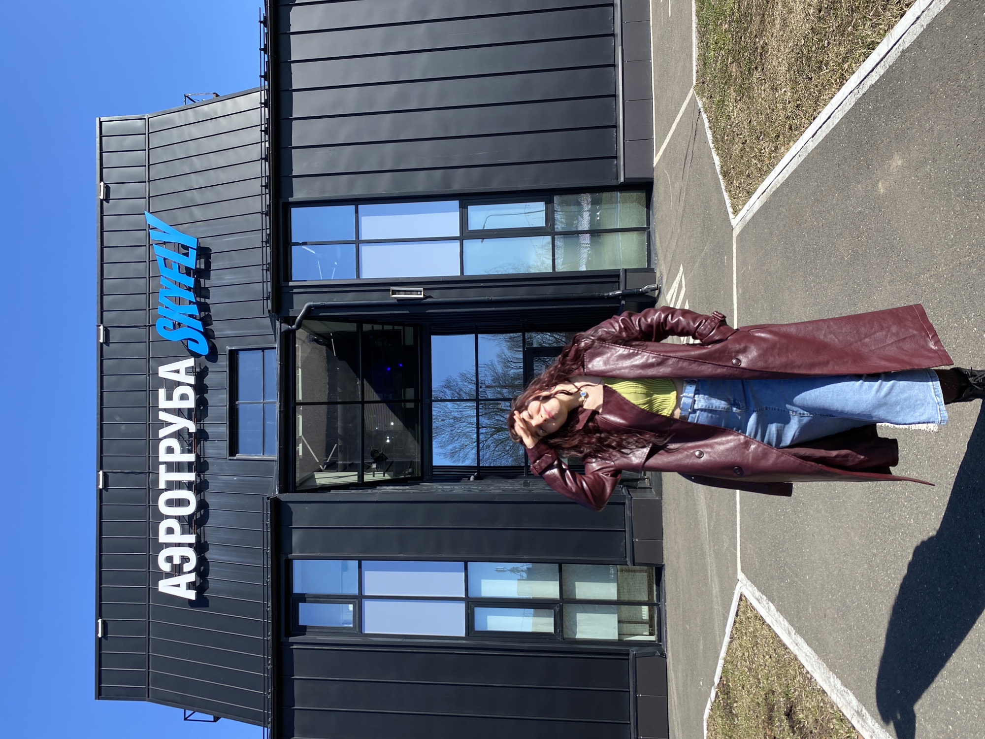 A full-body shot shows a woman in a burgundy leather trench coat, a light green shirt, and blue jeans, standing on a paved surface with grassy edges. She holds her hands up to her face. Behind her is a dark gray building with a facade of horizontal lines, windows, and a blue sign that reads "SKYFLY" and "АЭРОТРУБА." The location is Izhevsk, Russia. The sky is clear and blue.