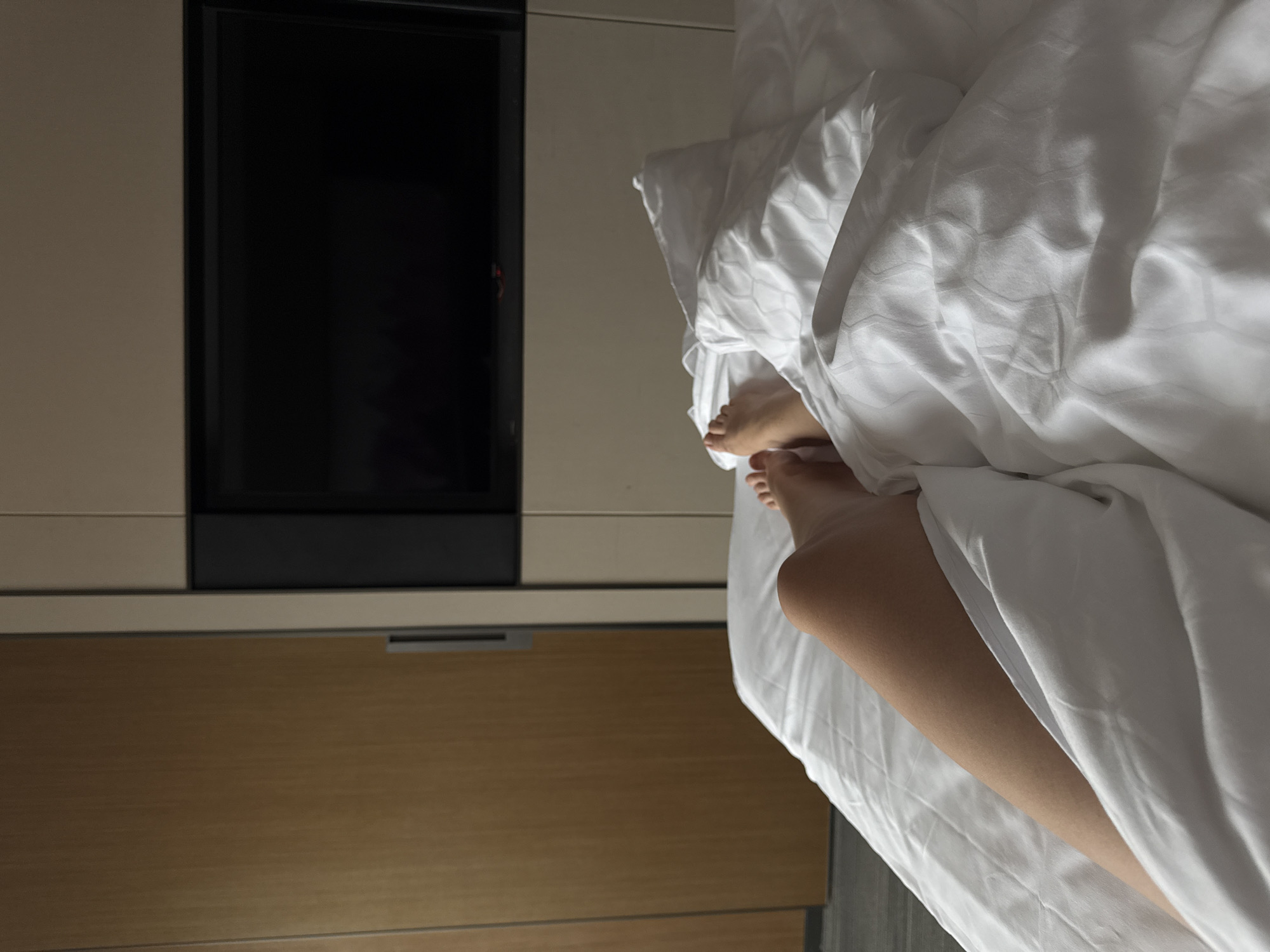 A bedroom view shows bare feet protruding from under white sheets on a bed. Beside the bed is a wooden bedside table, and a television set mounted on the wall above it. The exact location and the city of Zhangjiajie in China cannot be determined from the image alone.