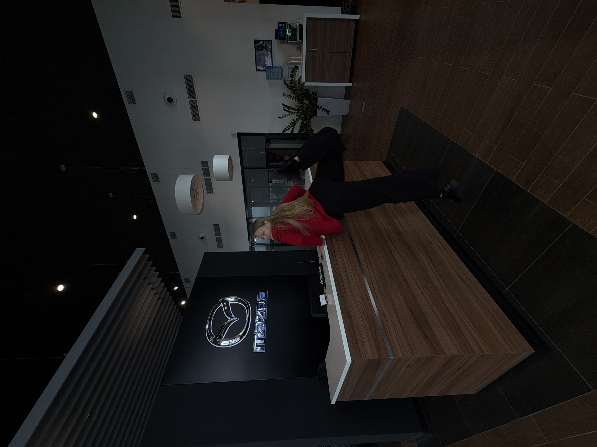 The image shows a person standing in the reception area of a Mazda dealership. The person has long blonde hair and is wearing a red sweater and black pants. They are standing behind a wooden reception desk with the Mazda logo prominently displayed on the wall behind. The background includes office furniture, plants, and lighting fixtures. The location is identified as Izhevsk, Russia.