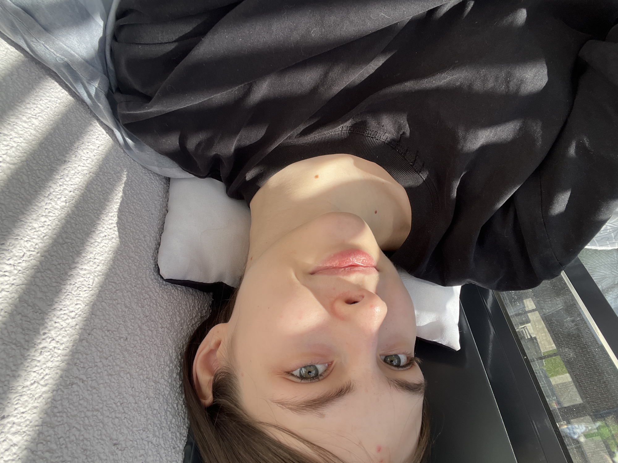A young woman lies on a bed in Saint Petersburg, Russia. Her head rests on a white pillow, with a black long-sleeve shirt covering her upper body. Sunlight streams in through a window, casting shadows across the scene. The room appears to be modern with views to the outside.