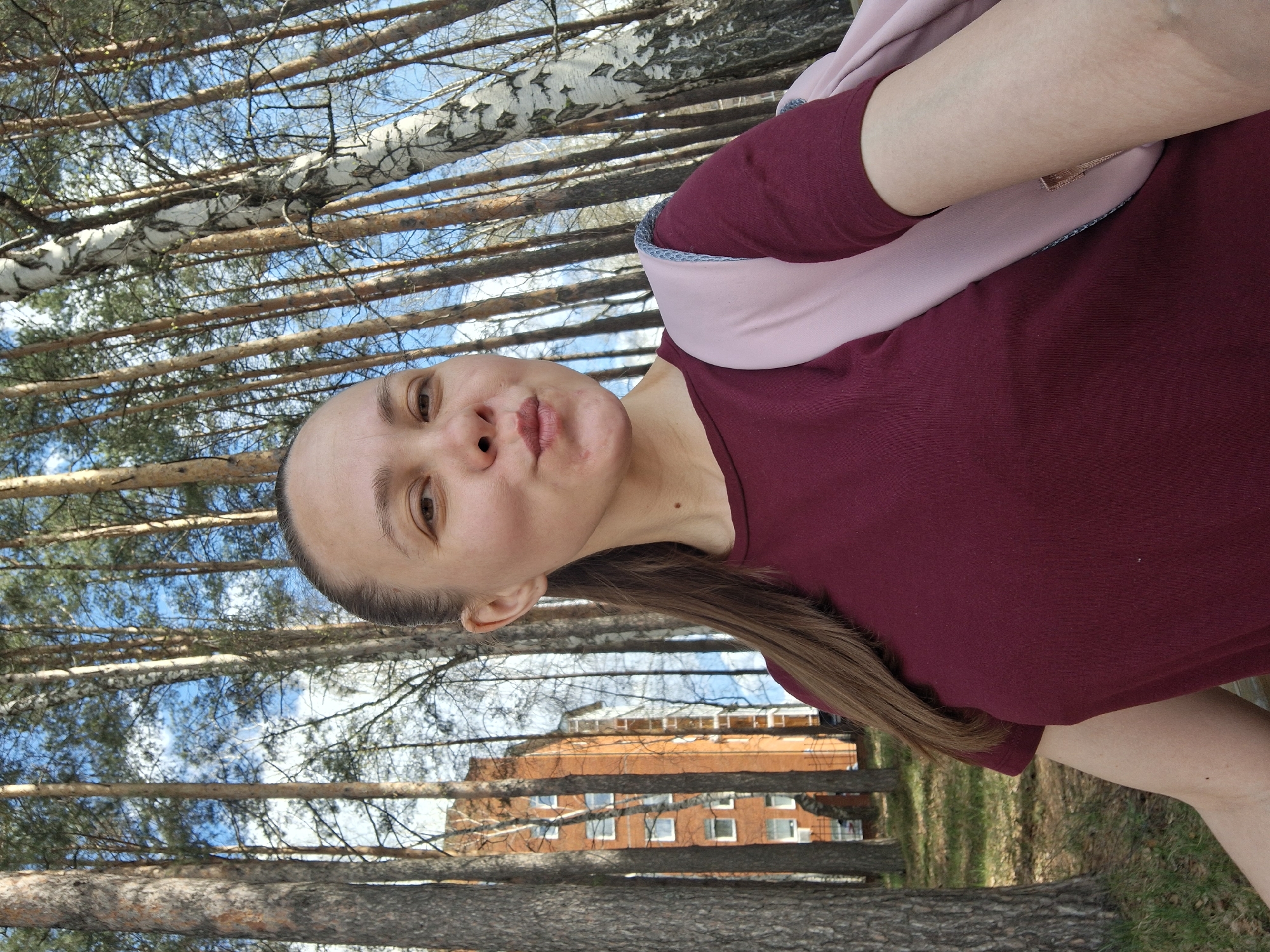 A woman poses for a selfie in a wooded area in Chaikovsky, Russia. She is wearing a maroon top and a light pink scarf. The background includes tall trees and a brick building.