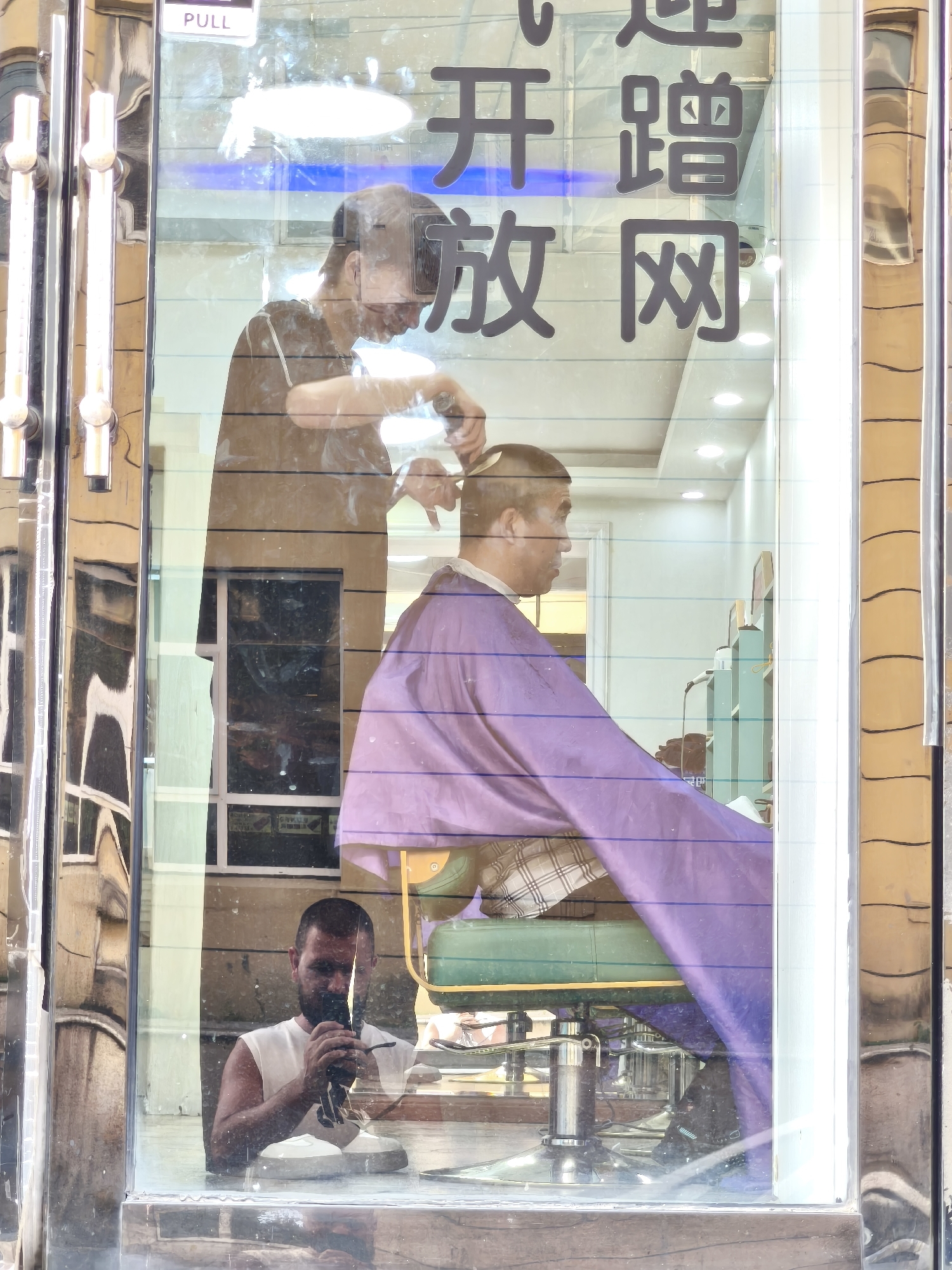 The image captures a reflection in a glass storefront, likely in Changchun City, China, as indicated by the Chinese characters visible on the glass. The reflection shows two men inside a barbershop. In the foreground of the reflection, a man wearing a white tank top is holding a phone and appears to be taking a selfie. He has a beard and short hair. Behind him, another man, presumably a barber, is cutting the hair of a customer. The barber is wearing a dark t-shirt and the customer is draped in a purple cape. The barbershop interior is visible, with a barber chair and some shelves in the background. The exterior of the storefront features chrome-plated pillars and a "PULL" sign. The reflections on the glass create a layered effect, making it difficult to distinguish between the interior and the exterior.