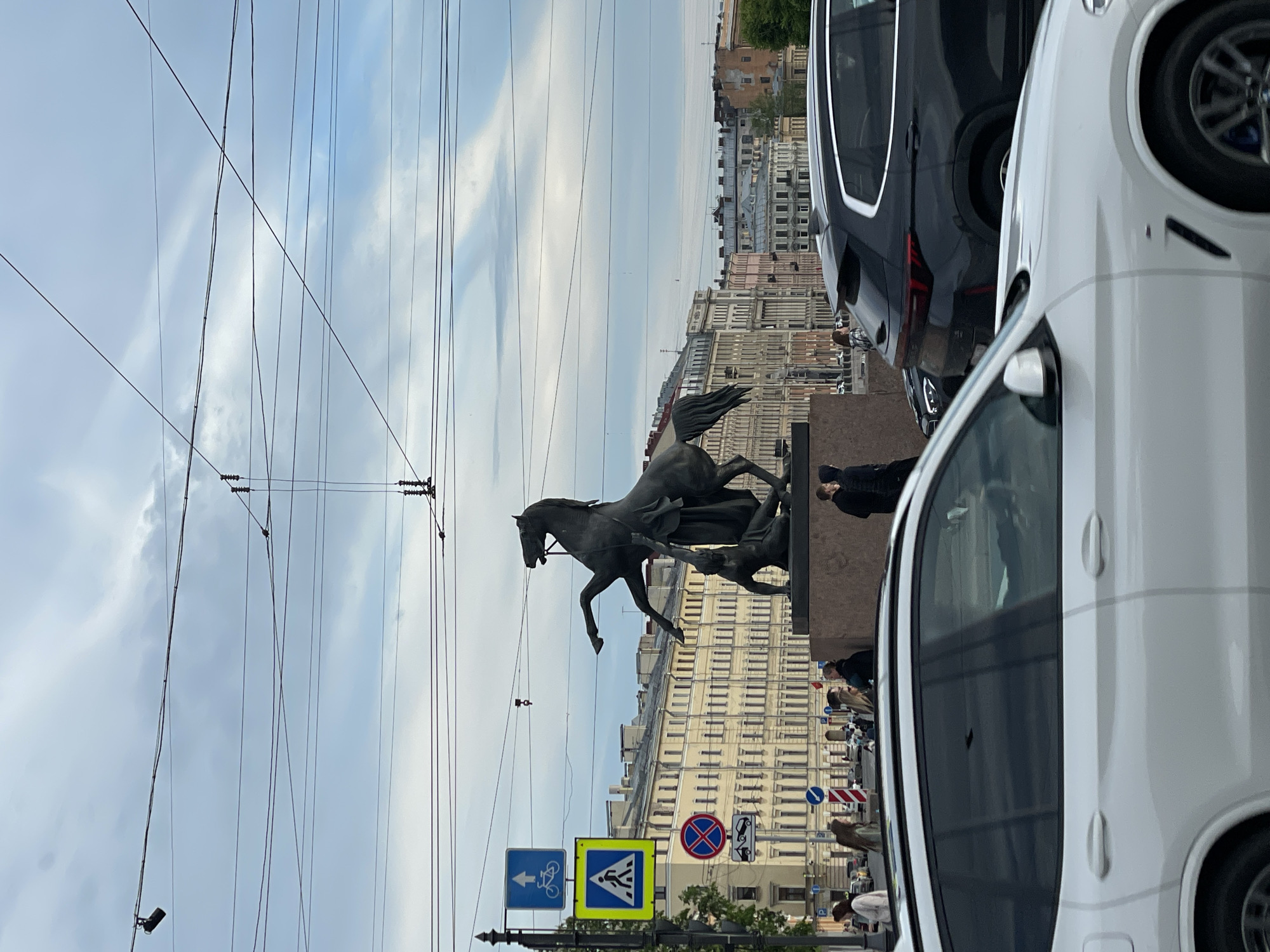 The image displays a lively street scene in Saint Petersburg, Russia, under an overcast sky during daytime. A prominent bronze equestrian statue, one of the Anichkov Bridge horses, stands in the mid-ground. It depicts a powerful rearing horse being controlled by a male figure, set upon a substantial stone base.
In the foreground on the right, a white SUV/crossover is partially visible, with a dark-colored SUV parked directly behind it. The sky above is crisscrossed with numerous overhead electrical wires, characteristic of a city with tram or trolleybus infrastructure.
Along the street, multi-story buildings with classical architectural elements and numerous windows line the background, reflecting the historical urban landscape of Saint Petersburg. Several pedestrians are scattered along the sidewalks and near the monument, engaging in routine city activities.
To the left of the monument, a cluster of road signs is visible: a blue rectangular sign with a white arrow pointing left and a bicycle symbol, a yellow square sign indicating a pedestrian crossing, a red circular "no stopping" sign, and a smaller white sign indicating a "tow-away zone." The scene captures a typical urban moment, blending historic artistry with modern city life.