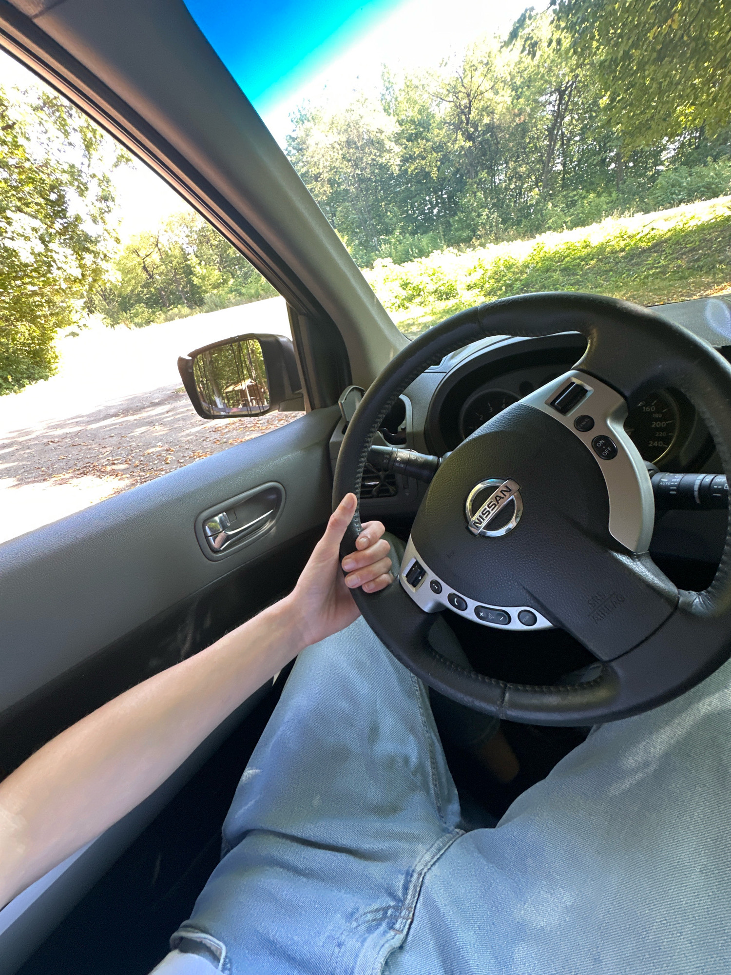 This is a first-person perspective image taken from inside a car, likely on a sunny day. The view is directed forward and slightly to the left, showing the interior of the driver's side of the vehicle and the road ahead. The steering wheel, with the Nissan logo prominently displayed, is in the foreground and right side of the frame. A person's arm, clad in light blue jeans, is resting on the steering wheel. The arm is bare from the elbow down, and the hand is lightly gripping the wheel.
Outside the car, a dirt road is visible, bordered by lush green trees and foliage. The sun is shining brightly, casting dappled light through the leaves. A side mirror is visible on the driver's side, reflecting a gazebo or small shelter in a wooded area. The sky is a clear, vibrant blue.
The image was taken in Shatava, Ukraine. The scene depicts a person driving on a rural road surrounded by nature, suggesting a leisurely drive through the countryside.