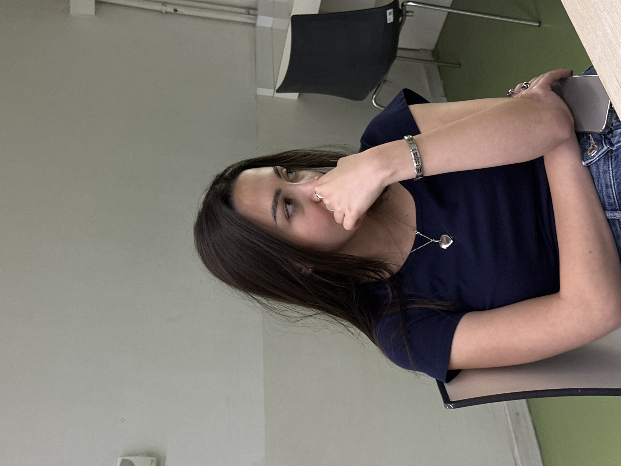 A young woman sits in a chair, wearing a navy blue shirt and denim jeans. She has dark hair and is wearing a silver bracelet and a silver necklace with a heart pendant. She is holding a phone in her left hand. She is likely indoors, judging by the walls and ceiling present in the background. There are also chairs present. There is no evidence to specify that it is in Yerevan, Armenia.
