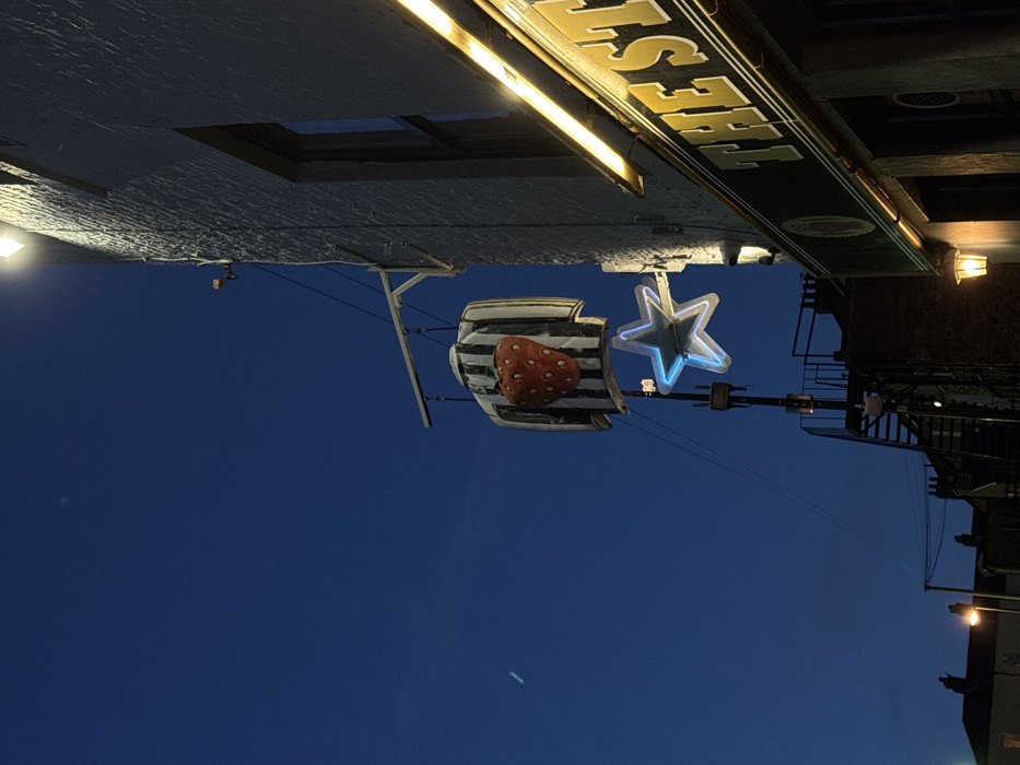 A rotated, nighttime image features a dark blue sky. In the foreground, a striped object with a large strawberry on it is suspended. Next to it is a glowing blue star. Above, the underside of a building is visible, illuminated by a light fixture. A sign reading "The Fest" is present, along with a set of stairs and additional lighting fixtures on the building.