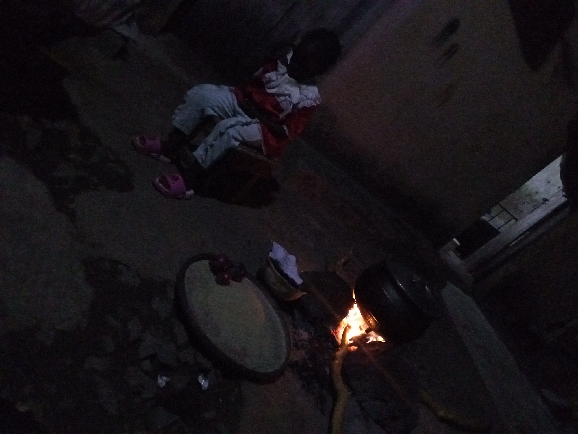 A dimly lit indoor space in Jos, Nigeria, features a child sitting on a low surface, wearing pink sandals, light-colored pants, and a red and white top. To the lower right, there is a small fire with a black pot resting on it. Adjacent to the fire is a circular tray containing red onions and a basket lined with a white cloth. The floor is uneven, and there are elements of what looks like a structure in the background.