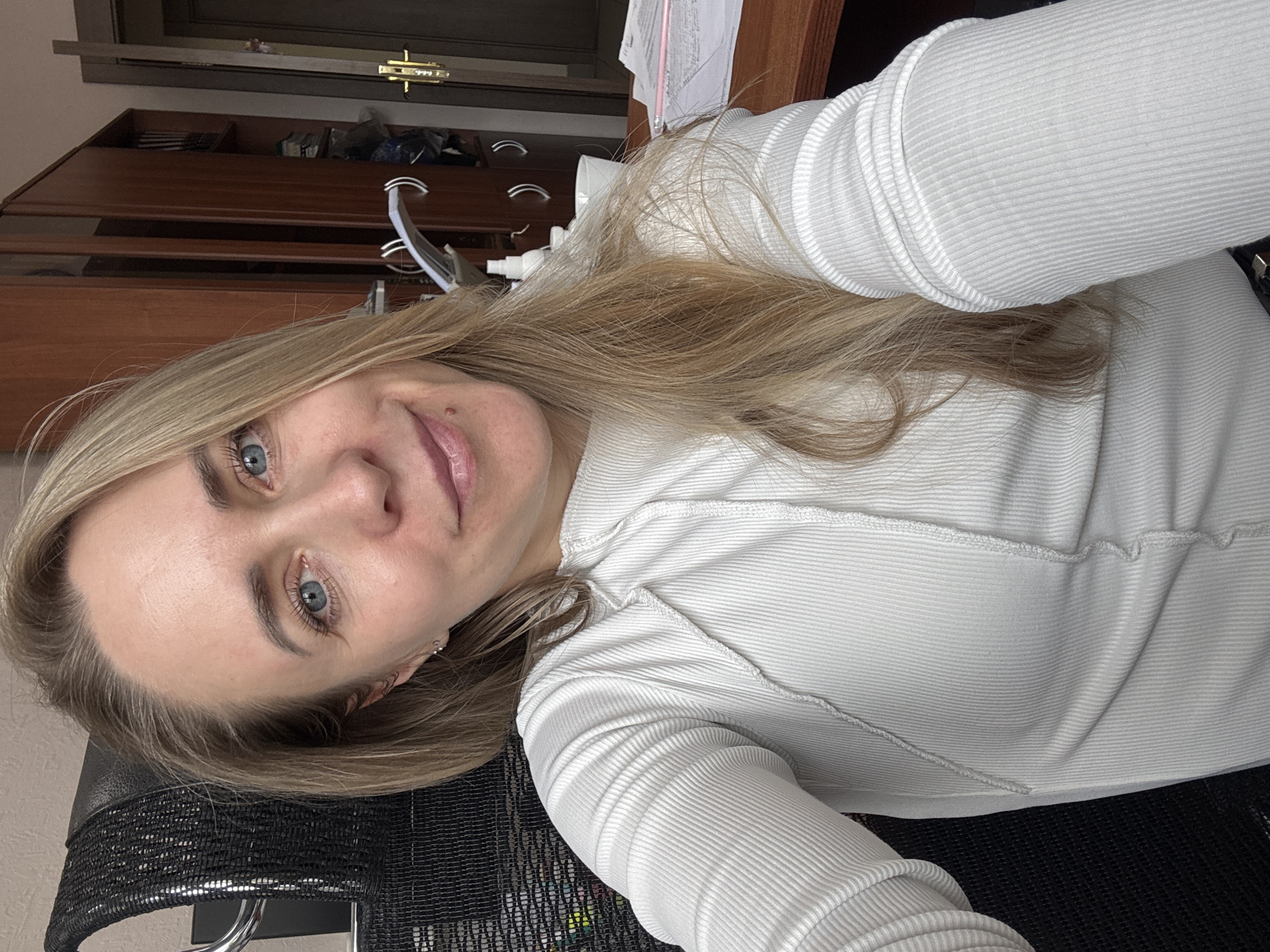 A blonde woman with blue eyes is in a chair in what appears to be an office. There is a wooden cabinet with drawers in the background. There is a computer monitor and paperwork on the desk. It is not possible to ascertain the location as Chaikovsky, Russia, without additional information.