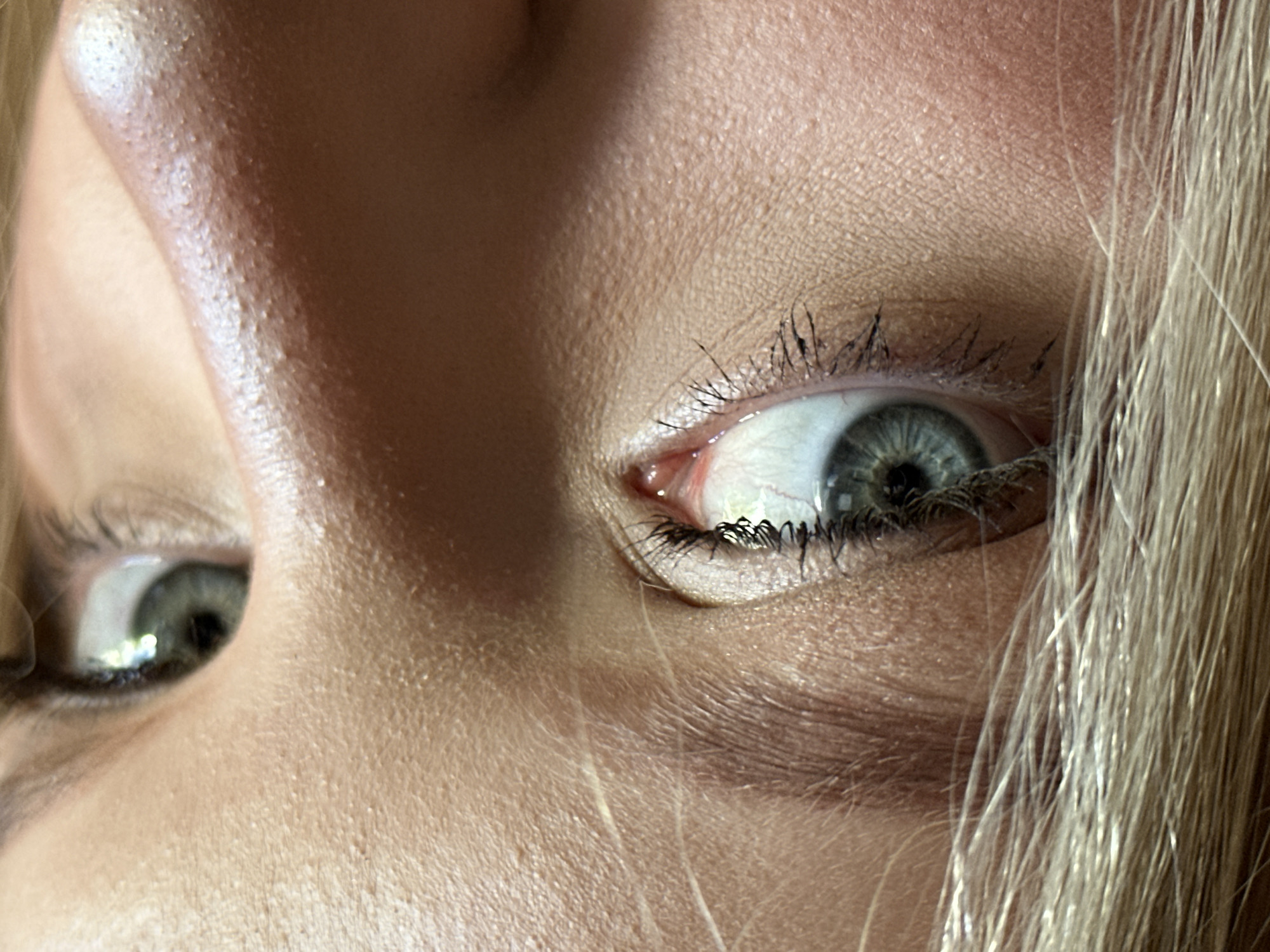 A close-up photograph shows a person's face from an extreme, almost inverted angle. The dominant feature is the individual's right eye, displaying a gray-green iris and dark lashes coated with mascara. The skin is fair and textured, reflecting light, particularly on the forehead. Strands of blonde hair are visible on the right side of the frame. No definitive connection to Chaikovsky, Russia, can be inferred.