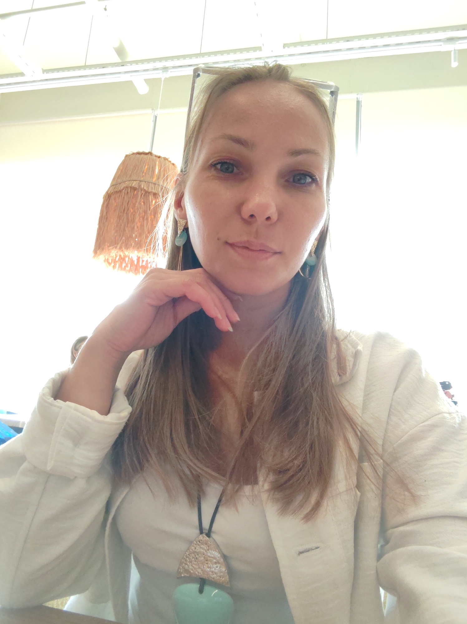 A young woman with blonde hair and blue eyes is taking a selfie. She is wearing a white blazer over a white top and a necklace with a large pendant. Her left hand is resting on her chin, and she is looking at the camera. She has earrings with light blue stones. In the background, there is a ceiling with fluorescent lights and a lampshade made of straw. The lighting is bright and washes out some details. The location is not specified in the image or any accompanying data, and therefore, cannot be determined to be Syktyvkar, Russia or any other specific city.