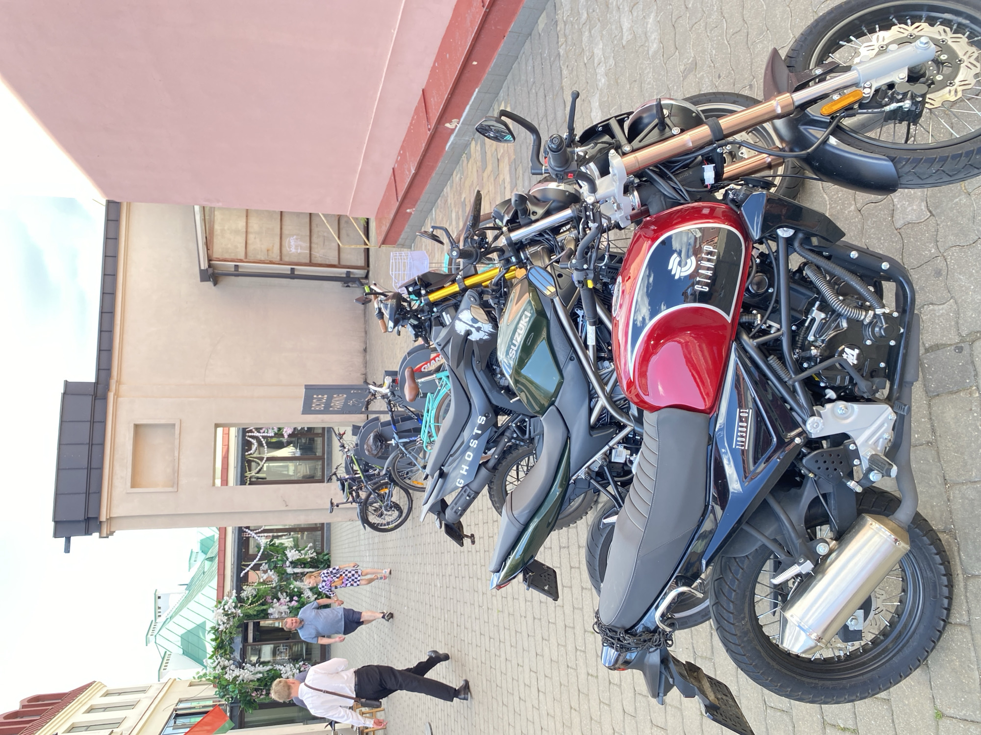 The image shows a row of parked motorcycles and bicycles on a cobblestone street, with a building facade in the background. Several people are visible, including a man in a blue t-shirt, a child in a checkered dress, and a person in a white shirt and black pants. The scene appears to be a public space in Minsk, Belarus, likely during the daytime.
The motorcycles are varied in style and color, with one prominent red motorcycle in the foreground featuring the text "OTTAKER" on its fuel tank. Other motorcycles are dark green and black. A "BICYCLE PARKING" sign is visible, indicating the area's purpose. The building facade is light-colored, with some windows and architectural details. The weather appears to be fair, with natural lighting suggesting a sunny day.
The people are casually dressed and appear to be walking or standing around the parked vehicles. The overall atmosphere seems relaxed and unhurried, consistent with a pleasant day outdoors. The presence of multiple motorcycles and bicycles suggests a mode of transportation popular in the area or perhaps an event or gathering related to these vehicles. The cobblestone street and building style provide clues about the location's urban environment.