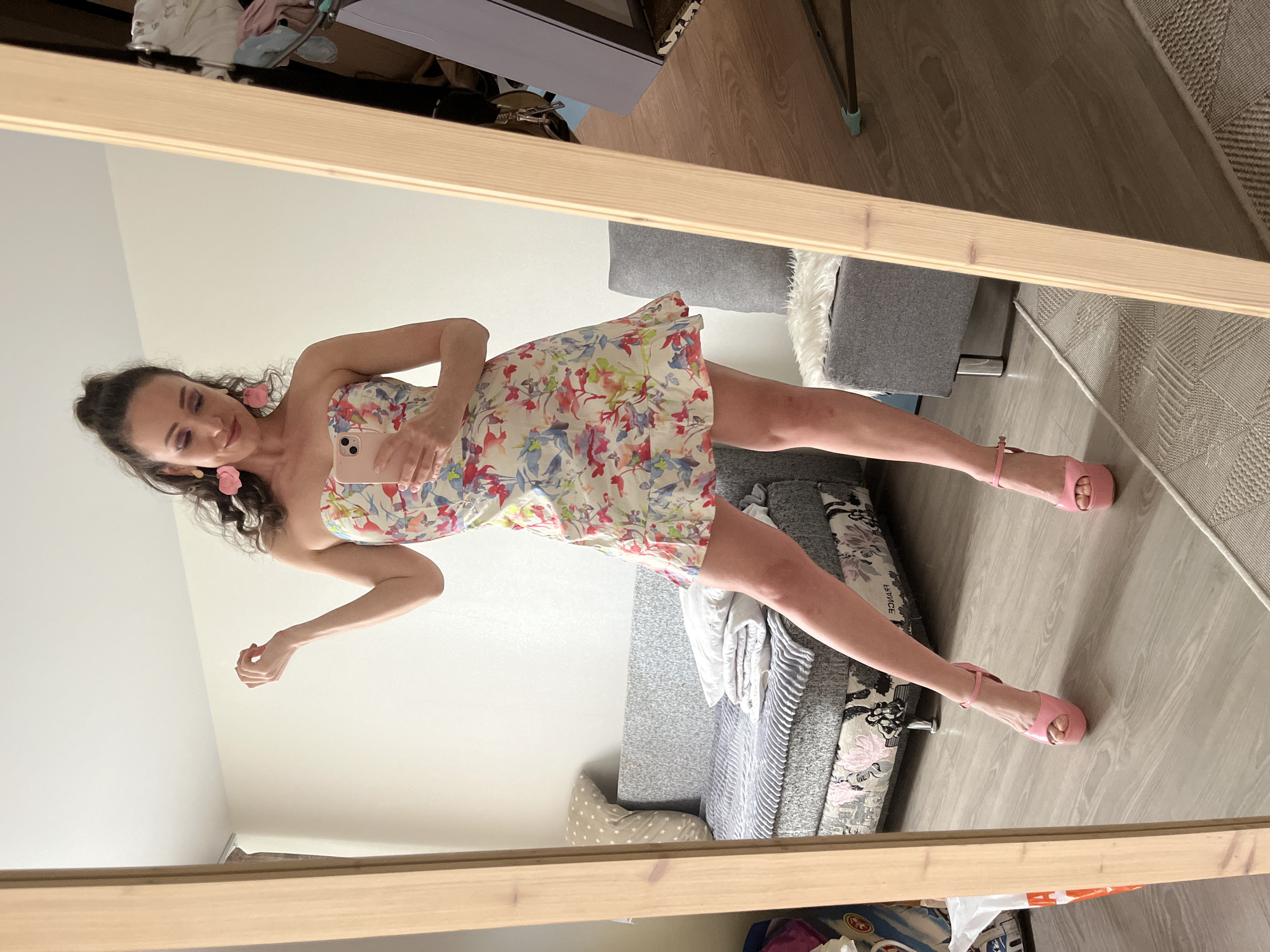 A woman stands in front of a full-length mirror, taking a selfie. She is wearing a strapless, floral-print mini dress and pink platform heels. Her dark hair is pulled up in a messy bun, and she has large pink flower earrings. In the background, a grey sofa with white pillows and a grey chair with a white furry rug are visible. The woman's left hand is raised, and she is holding her phone in her right hand. The room appears to be a bedroom or living room, with wooden floors and white walls. There is no specific indication of the location being in Kazan, Russia.