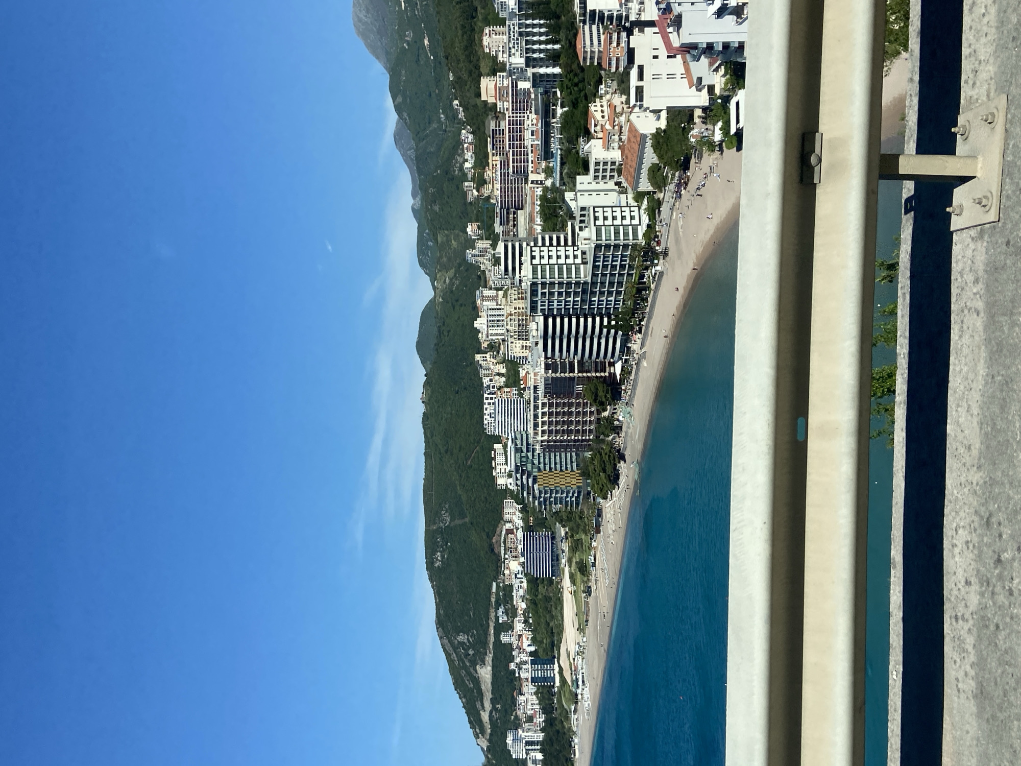 This image captures a panoramic view of a vibrant coastal town from an elevated perspective, looking down onto a sandy beach and the Adriatic Sea. The bright blue sky, with a few faint wispy clouds, fills the upper left portion of the frame (when correctly oriented).
In the mid-ground, a long, sandy beach stretches along the coast, populated by numerous small figures of people, suggesting leisurely activities such as sunbathing or walking. The calm, clear sea transitions from a light turquoise hue near the shore to a deeper blue further out, inviting swimmers.
Behind the beach, a dense urban landscape unfolds. A mix of modern, multi-story buildings, likely hotels and resorts with distinctive architectural designs, lines the coastline. Some structures feature prominent balconies and varied facades, while others exhibit more traditional elements, including red-tiled roofs, as they extend up the lower slopes of the mountains.
The background is dominated by a majestic chain of verdant mountains, thickly covered in green trees and vegetation, forming a dramatic natural backdrop to the developed area.
In the foreground, on the right side, a sturdy, light-colored guardrail or barrier with visible bolts and structural supports indicates the elevated viewpoint, suggesting the photo was taken from a road, bridge, or high vantage point.
The scene is bathed in bright, clear sunlight, implying a warm, sunny day, likely during midday or early afternoon given the strong shadows. The overall atmosphere is one of a bustling summer resort. This location is confirmed as Budva, Montenegro. No visible text is present in the image.
