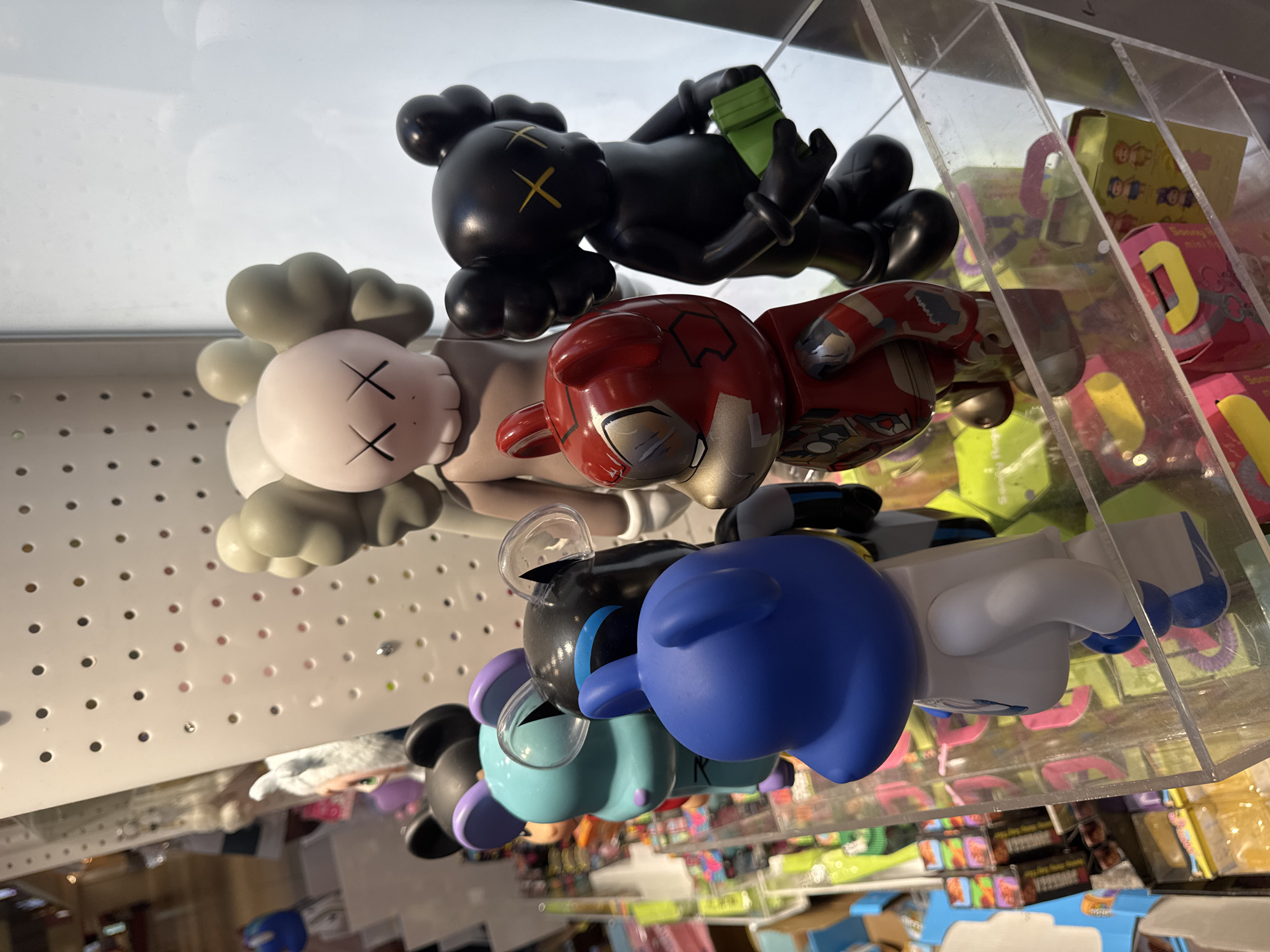 This media file presents a close-up, upward-angled view of collectible vinyl art toys displayed in what appears to be a retail store or gallery in Rosemont, United States of America. The image, originally rotated 90 degrees clockwise, showcases multiple figures arranged on clear acrylic shelves.

At the top of the corrected image, a light greyish-tan figure with a pinkish-white face and distinctive black "X" eyes, characteristic of KAWS Companion designs, is prominent. Below it, a black KAWS-style figure with yellow "X" eyes lies horizontally, holding a bright green rectangular object. Further down the display, a red and silver figure with a distressed, metallic appearance, possibly a stylized robot or superhero, is visible.

The lower part of the arrangement includes a black and blue figure with blue ears, followed by a light blue figure with purple ears. At the very bottom, a figure with a large blue head, white body, and blue shoes completes the immediate display.

The figures are set against a background featuring a white pegboard surface. Behind and to the right of the figures, various colorful, blurred boxes of merchandise are visible through clear dividers, suggesting a busy retail environment. Some legible text on these boxes includes "Boney mini box," "OOF," "101," and "MYSTERY." The overall bright lighting indicates an indoor setting, likely during the daytime. No people are visible, and the scene focuses entirely on the static display of the art toys.