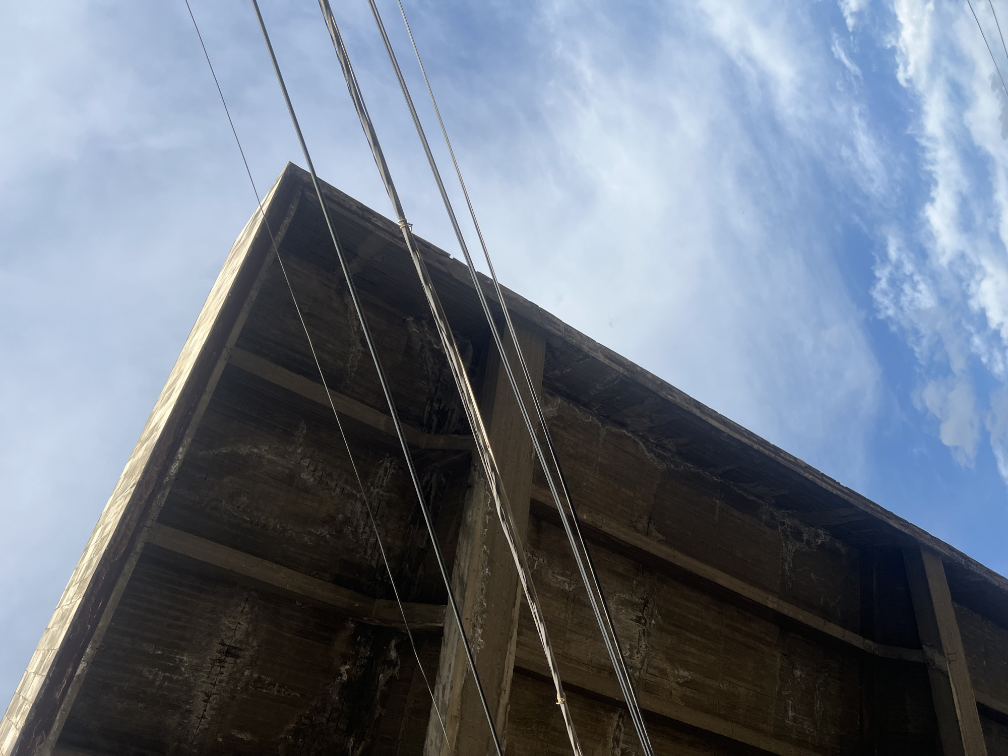 The image depicts an upward-looking view beneath a large, weathered concrete overhead structure, likely a bridge or overpass, set against a partly cloudy sky. The concrete structure occupies the upper left and extends diagonally downwards towards the lower left. Its underside exhibits a textured, dark grey-brown surface with visible parallel support beams and lighter patches of efflorescence or staining. Several utility cables, appearing as dark horizontal lines, traverse the frame between the concrete structure and the sky. One cable in the lower center shows a minor knot or wrap around it. The sky, visible in the upper right and lower right sections, is a clear blue with dispersed white cumulus and cirrus clouds. The lighting suggests daytime conditions. The specific location within the city of Yerevan, Armenia, is not discernible from the provided visual data.