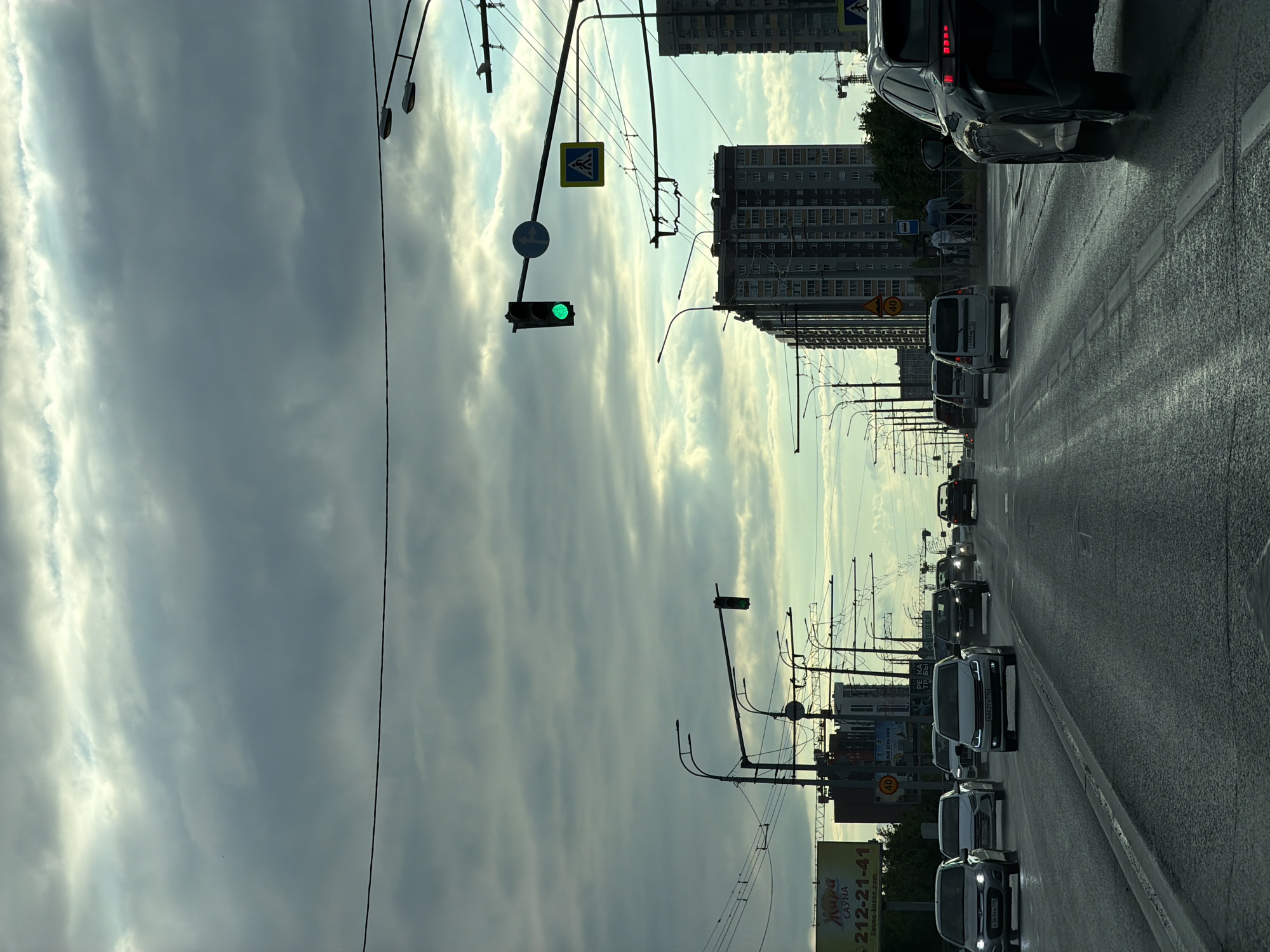 This is a tilted, eye-level, outdoor photograph taken from a vehicle traveling down a wet, multi-lane road lined with buildings and power lines. The sky is overcast with dramatic clouds. Traffic is moderate, with several cars visible in front and behind the camera. The scene appears to be in a city, possibly during the daytime given the natural light, though the heavy cloud cover suggests a potentially dim or approaching storm.
In the foreground, we see the road surface, which is wet and reflective, indicating recent rain. Several cars are lined up, their headlights and taillights visible. Further down the road, there are more vehicles, including what appears to be a bus. Tall, modern buildings with many windows line the right side of the road, suggesting an urban environment. Power lines and street lights are prominent features, crisscrossing the sky and road.
On the left side of the road, there are traffic signals, including one with a green light illuminated. A pedestrian crossing sign is also visible. In the distance, construction cranes are visible, hinting at ongoing development.
There is some visible text on a billboard on the right side of the road. The text includes Russian words like "Жара" (Heat) and "САУНА" (Sauna), along with a phone number "212-21-41". Another sign has "40" on it, likely indicating a speed limit. The overall mood of the image is somewhat somber due to the weather, but also conveys a sense of movement and urban life.