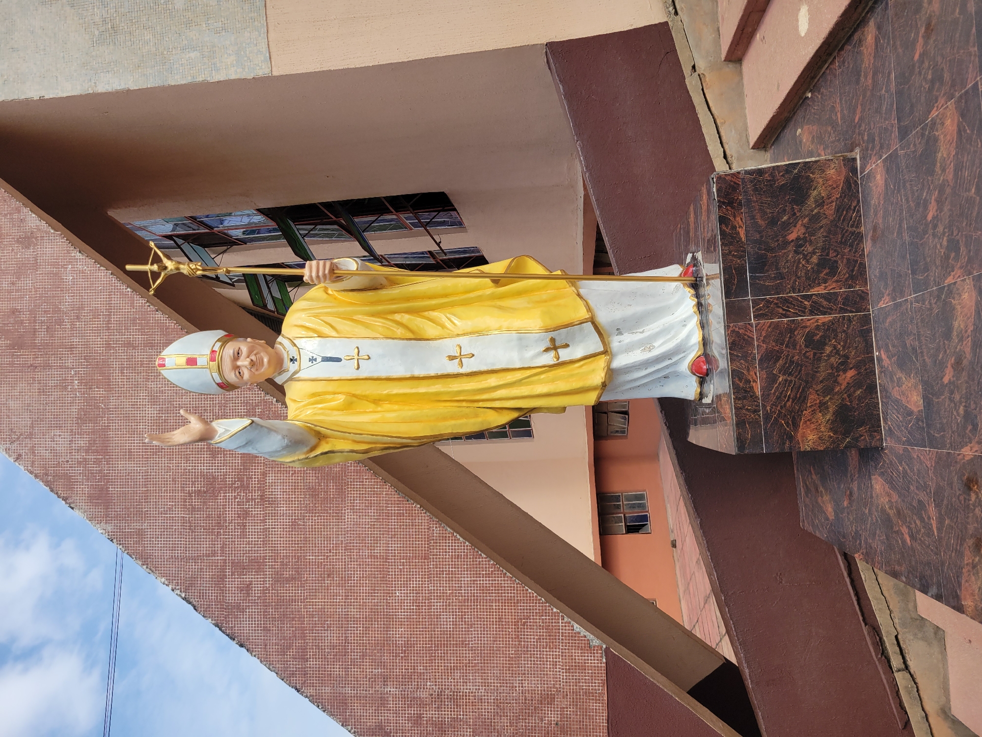 A full-body statue of Pope John Paul II stands on a patterned marble pedestal outside a building with a large, angled, tiled wall and stained-glass windows. The statue depicts the Pope in full regalia, wearing a white and yellow papal vestment with golden crosses, a papal tiara, and holding a golden crucifix on a staff in his right hand. His left hand is raised in a wave. The building has a pink-orange facade with visible windows. The ground area is paved with tiles and there are stone steps leading to the right. The sky above is blue with scattered clouds.