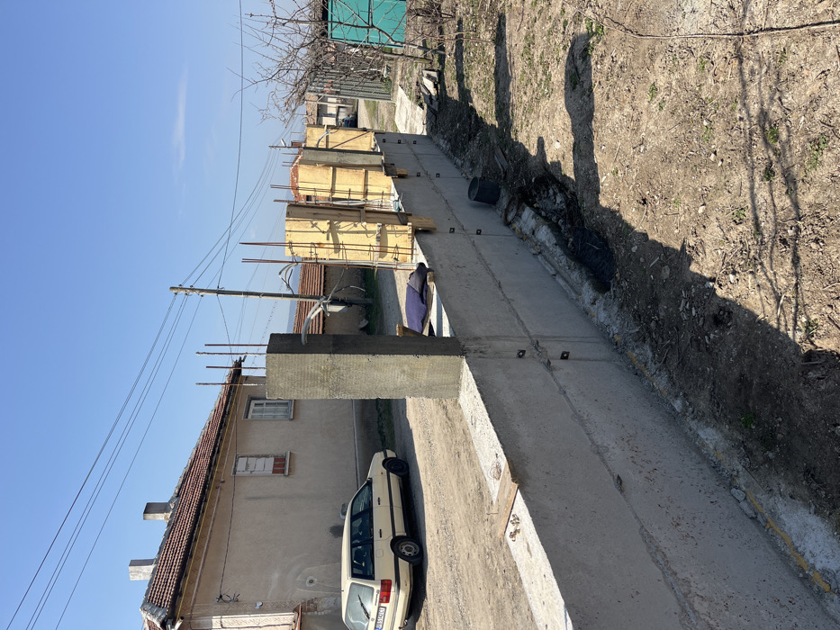The image depicts an outdoor construction scene in Zlatovrah, Bulgaria, under a clear blue sky. In the foreground, a concrete wall extends diagonally from the lower-left to the mid-right. Vertical steel reinforcing bars protrude from the top of the concrete wall at regular intervals. Several sections of wooden formwork, containing vertical rebar, are erected along the wall, prepared for pouring additional concrete columns. One finished concrete column stands upright on the left portion of the wall. A narrow trench, containing excavated dirt and dark plastic sheeting, runs along the base of the wall on the viewer's side. To the left of the concrete wall, a light-colored station wagon, bearing Bulgarian license plate "PB 1424 CT," is parked on a dirt surface. Behind the vehicle, a two-story, light-colored residential building with a red-tiled roof and two chimneys is visible; one window on this building is boarded. Overhead power lines span the upper midground, supported by a wooden utility pole. The background shows additional distant buildings and a clear horizon with faint hills. No individuals are present in the scene.
