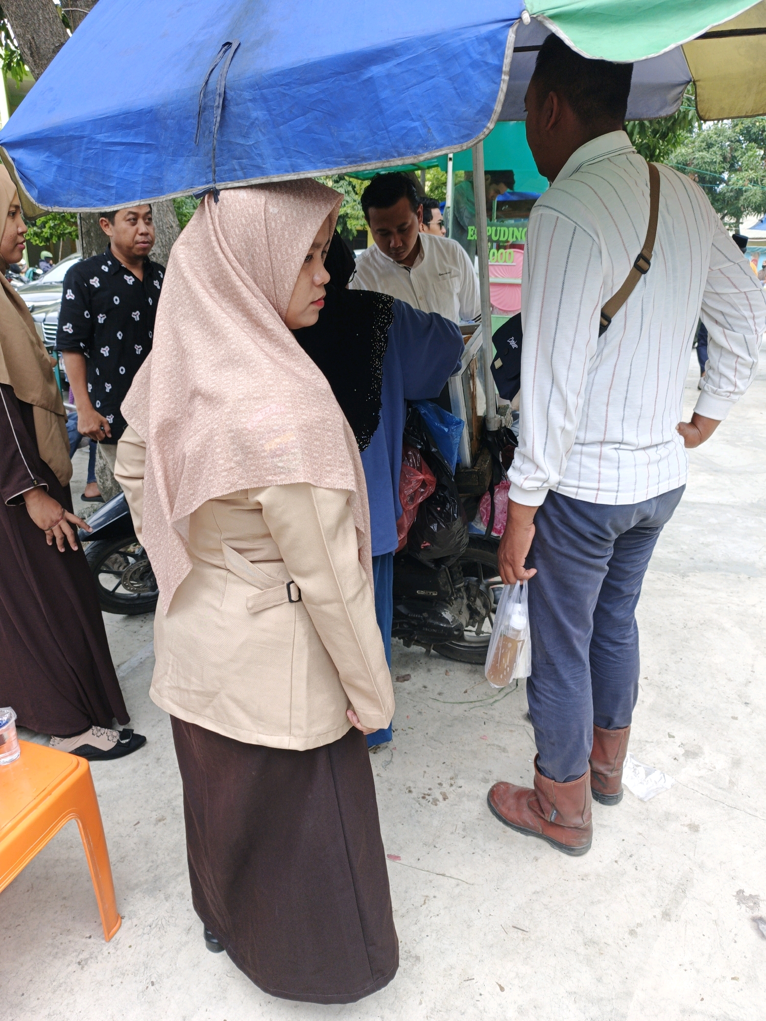 In Manunggal, Indonesia, a group of individuals are gathered under a blue and green tarp, likely at a street stall. A woman in the foreground, wearing a tan hijab and a matching jacket over a dark skirt, is looking to her right. To her left, another woman in a brown hijab and dress stands near an orange plastic table. Behind them, a man in a black patterned shirt looks towards the left. Further back, a man in a white striped shirt with blue and red lines is holding a plastic bag with a drink. He is wearing dark blue pants and brown boots. A motorcycle is parked behind the stall. The ground is paved with concrete.