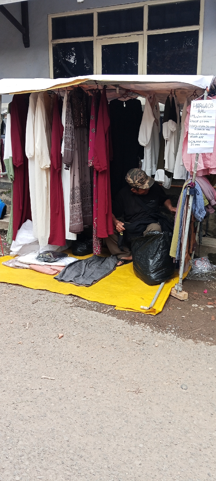 A man is sitting on a yellow mat in front of a stall with clothes hanging from a canopy. The stall is located in front of a building with a large window. The man is wearing a black shirt and black pants. He is looking at the ground. There is a black garbage bag next to him. The clothes hanging from the canopy are mostly long dresses and shirts, and there are some towels hanging on a rack behind the man. There is a sign hanging from the canopy that says "Harga 08" and lists prices for different types of clothing. The stall is located in front of a building in the city of Kutawaringin, Indonesia, at the coordinates -6.974 latitude and 107.519 longitude. The ground is covered in gravel and dirt. The building is painted a light blue color. The window in the building is made of glass and metal.