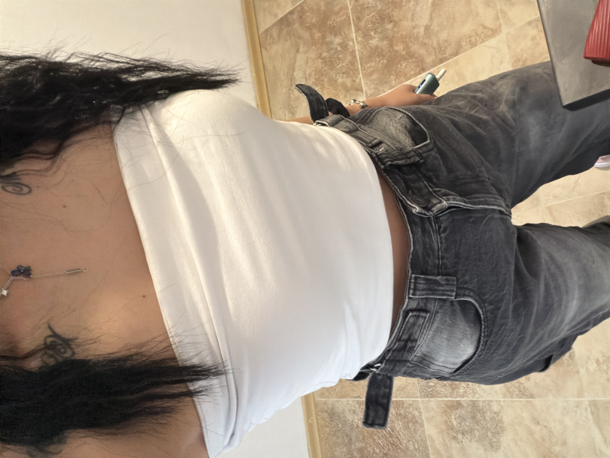 An undated, medium-shot, waist-up, indoor photograph shows a person from the chest down, wearing a white strapless top and distressed dark wash jeans. The person has long, dark hair. A silver necklace with a small, purple pendant is visible on the chest. The background consists of tiled flooring and a plain wall. The person is holding a small, green electronic device with a white tip in their right hand. The scene does not provide any specific clues that would allow for pinpointing a location within the city of Plovdiv, Bulgaria.
