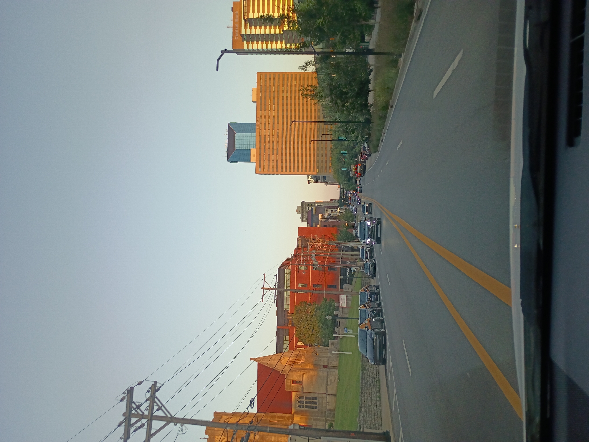 The image is a street-level view of a city street in Lexington, United States of America, likely taken from inside a vehicle. The sky is a clear, pale blue, suggesting it is either early morning or late afternoon. On the left side of the road, there is a large, red-brick building with a stone foundation, resembling a church, with Gothic-style windows. A few cars are parked along the left curb, and a traffic light is visible. Further down the street on the left, more red-brick buildings are present, some with modern glass facades.
On the right side of the road, there are several trees and a sidewalk area with a modern street lamp. In the distance, a line of cars is traveling down the street. Prominently visible are several tall buildings. One is a large, orange-tinted building with many horizontal windows, identified as the Hilton. Above it, a smaller, blue-glass-covered building stands out. To the right of the Hilton, another tall building, illuminated by the setting or rising sun, displays the "Central Bank" sign. The street itself is paved asphalt with double yellow lines in the center, indicating lanes of traffic. The overall scene depicts a typical urban environment with a mix of historic and modern architecture, alongside the presence of vehicular traffic.