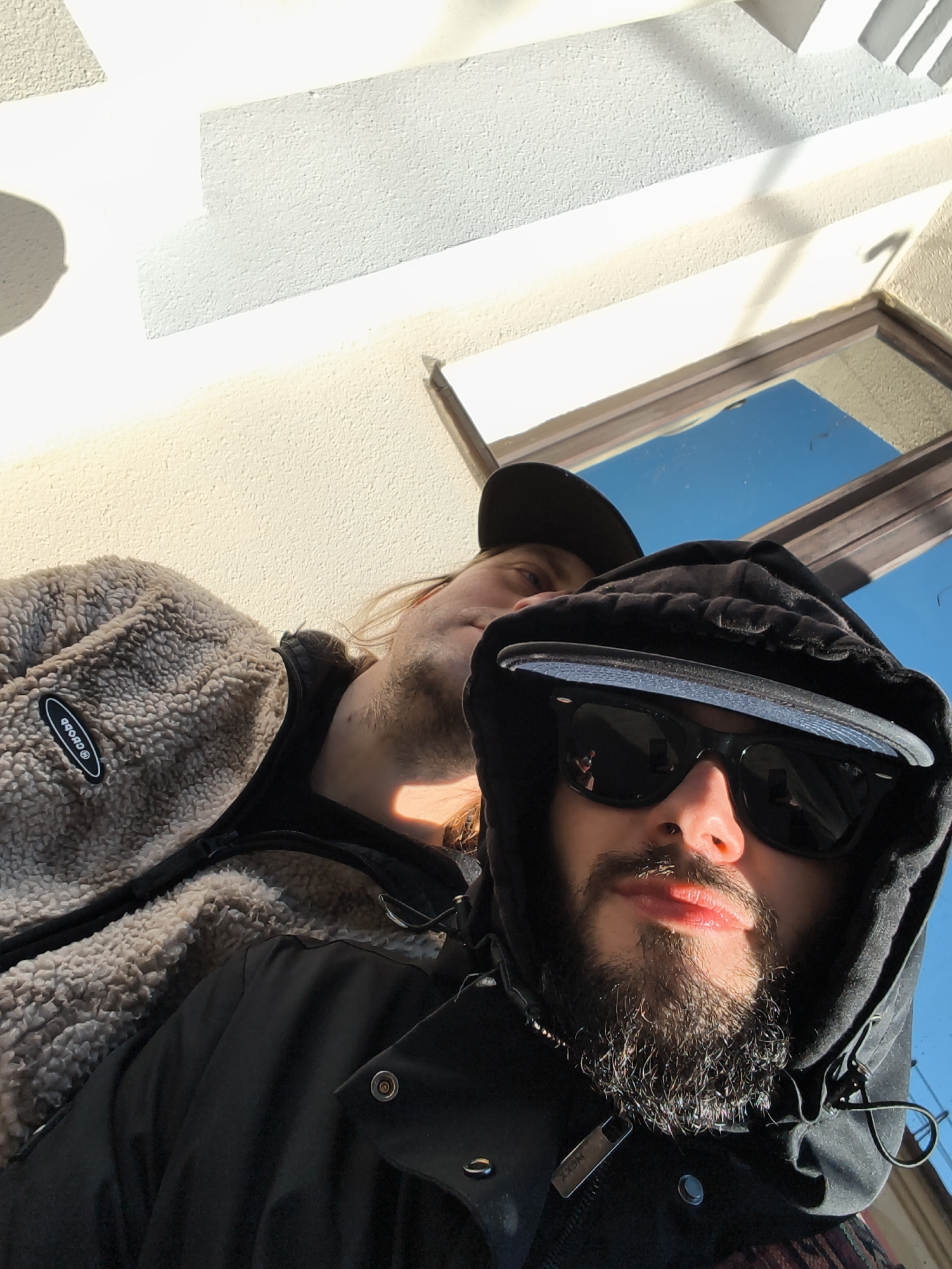 Two men are in the image. The man in the foreground wears a black hooded jacket, sunglasses, and a baseball cap. He has a beard and mustache. The second man is behind him, wearing a tan sherpa jacket and a baseball cap. They are outside with a building facade visible in the background. The building has a white wall and a window reflecting a blue sky. The location could be Hrodna, Belarus.