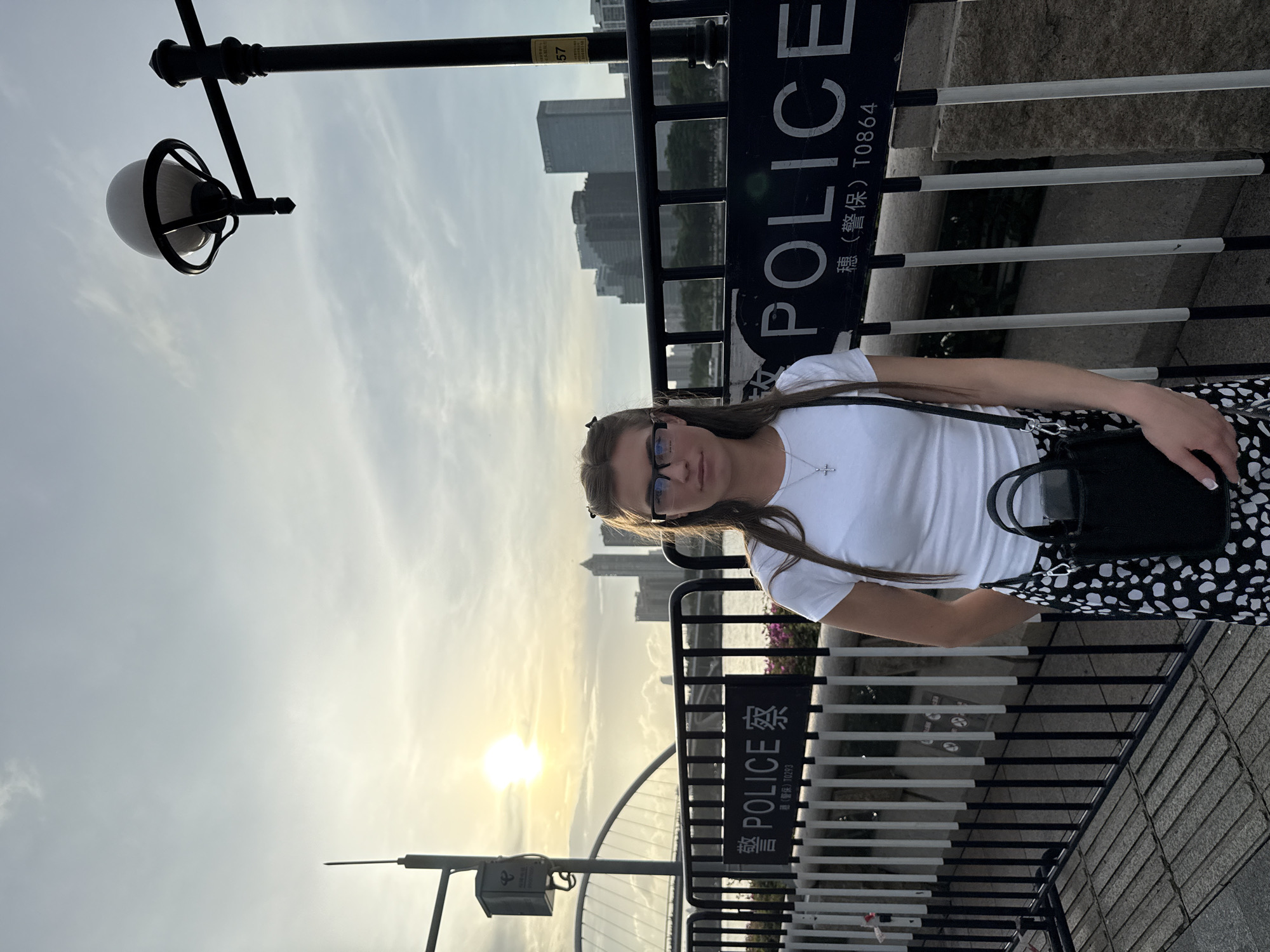 A young Caucasian woman stands in front of a black metal barrier with "POLICE" written in English and Chinese characters. She is wearing glasses, a white t-shirt, and a black and white patterned skirt. She has a black purse slung over her shoulder. The sun is setting behind a cityscape of tall buildings and a bridge. The scene appears to be in Haizhu District, China, likely near a waterfront area. The lighting suggests it is late afternoon or early evening. The barrier is set up along a paved walkway, and there are streetlights visible in the background. The overall impression is of a tourist or local pausing for a photograph in an urban environment.