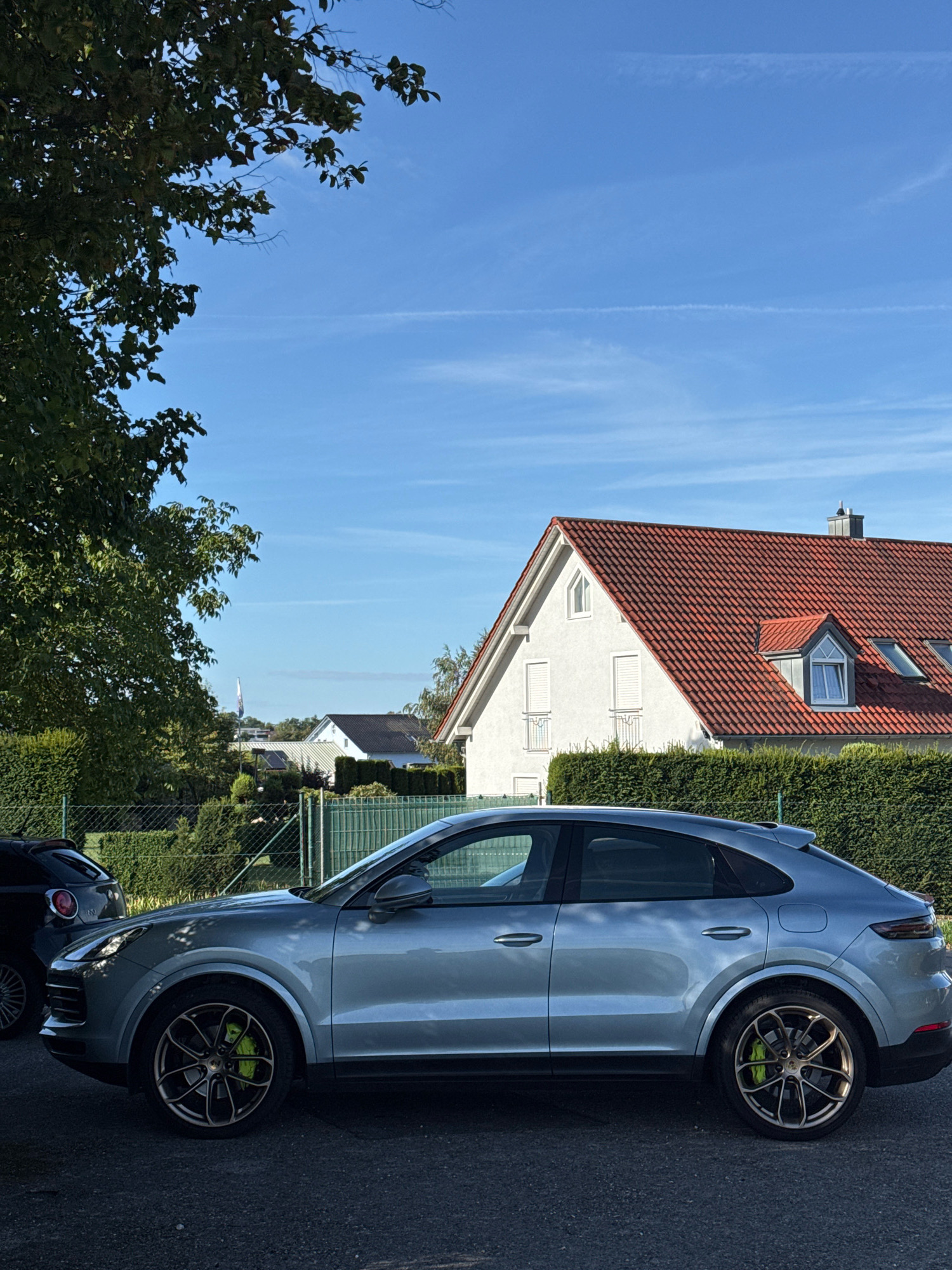 The image depicts a light blue Porsche Cayenne coupe parked on an asphalt road in Wettstetten, Germany. The car is viewed from the side, showcasing its sleek design and contrasting bright lime-green brake calipers. Behind the car, a dense, well-maintained green hedge forms a boundary. Further back, a white house with a prominent red-tiled roof and dormer windows is visible, set against a clear blue sky with faint contrails. To the left of the house, a tall tree with dark green foliage overhangs the scene. A dark-colored compact car is partially visible to the left of the Porsche. The overall environment suggests a residential area with manicured landscaping and clear weather.
